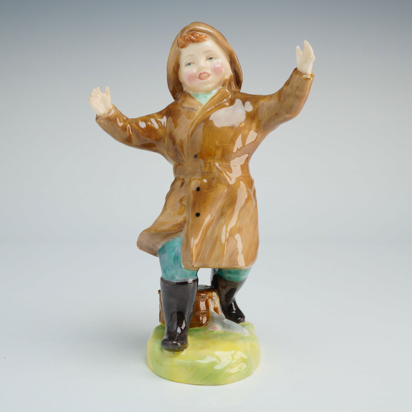 Royal Doulton Figurine, The One That Got Away HN2153: The figure depicts a young boy in fishing attire, wearing a glossy brown hooded raincoat, teal trousers, and black waders. He is posed with arms outstretched, standing on a base decorated in green and