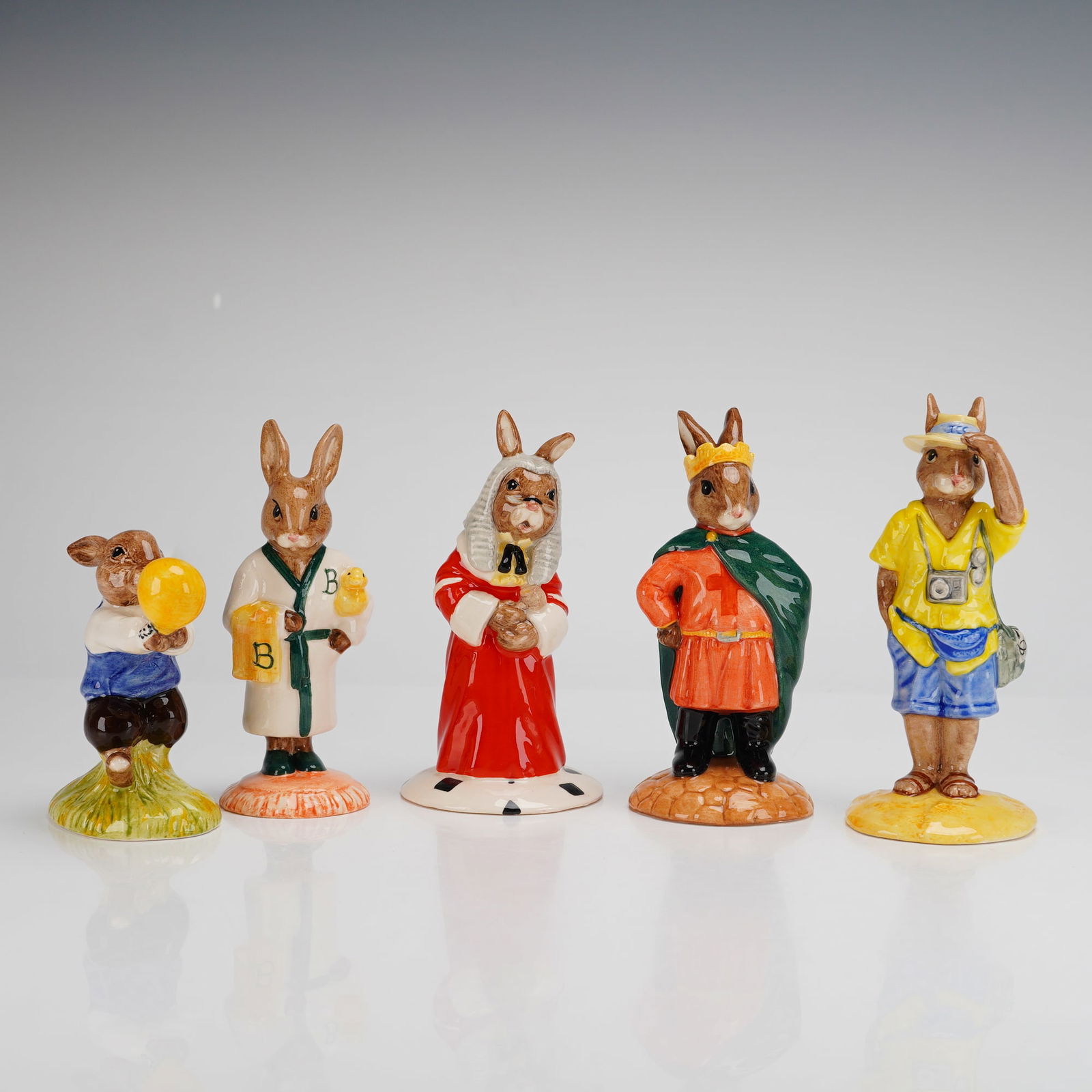 5pc Royal Doulton Bunnykins Figurines Bathtime Tourist Prince John Judge: A set of five Royal Doulton Bunnykins figurines crafted in hand-painted glazed ceramic. The collection includes the following characters: Harry with a yellow balloon (DB73), Bathtime depicted in a rob