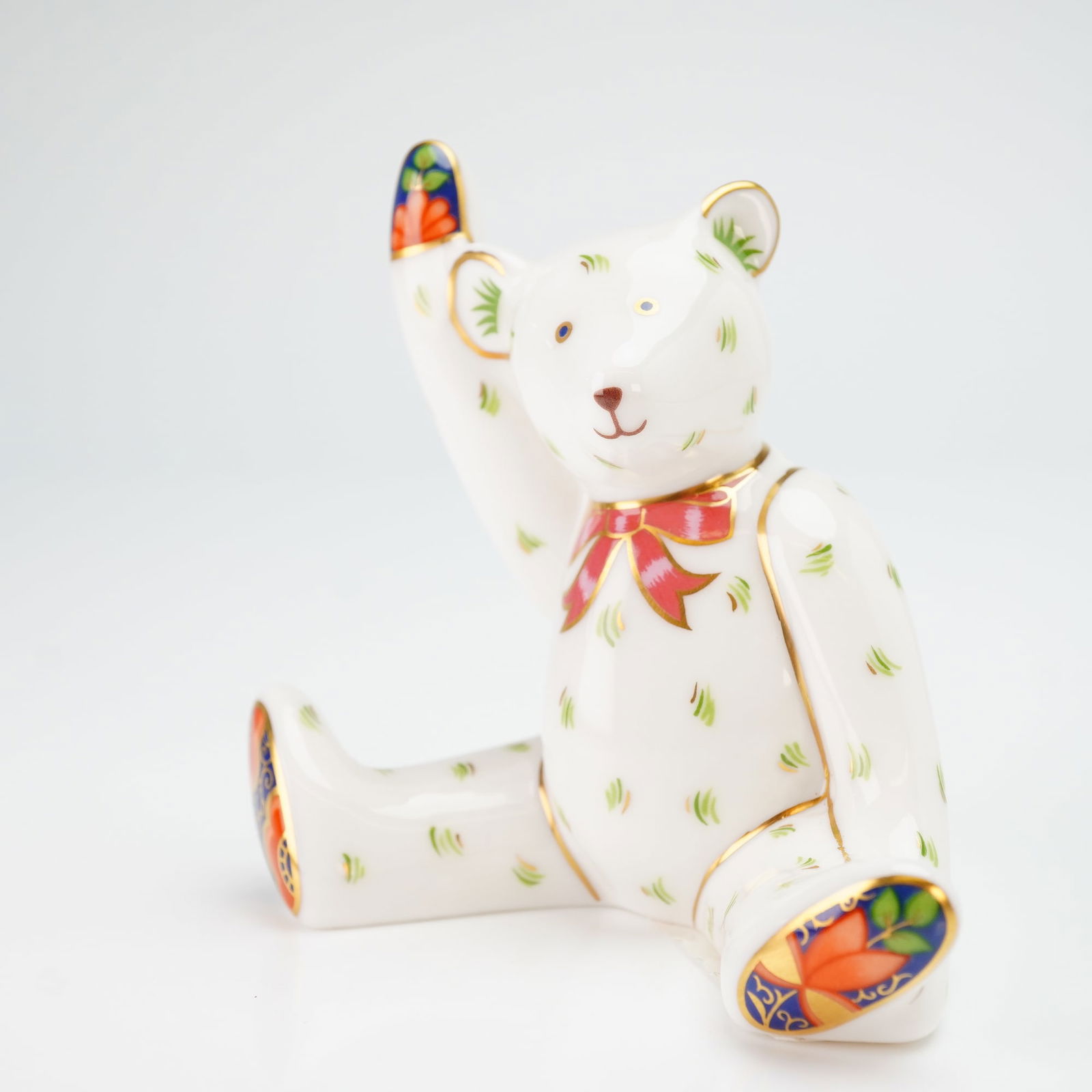 Royal Crown Derby Figurine, Mini Victoria Waving Teddy Bear: The figurine is modeled as a seated teddy bear with its right arm raised in a waving gesture. The piece is decorated with green sprigs over a white ground, featuring a red and gold bow around its