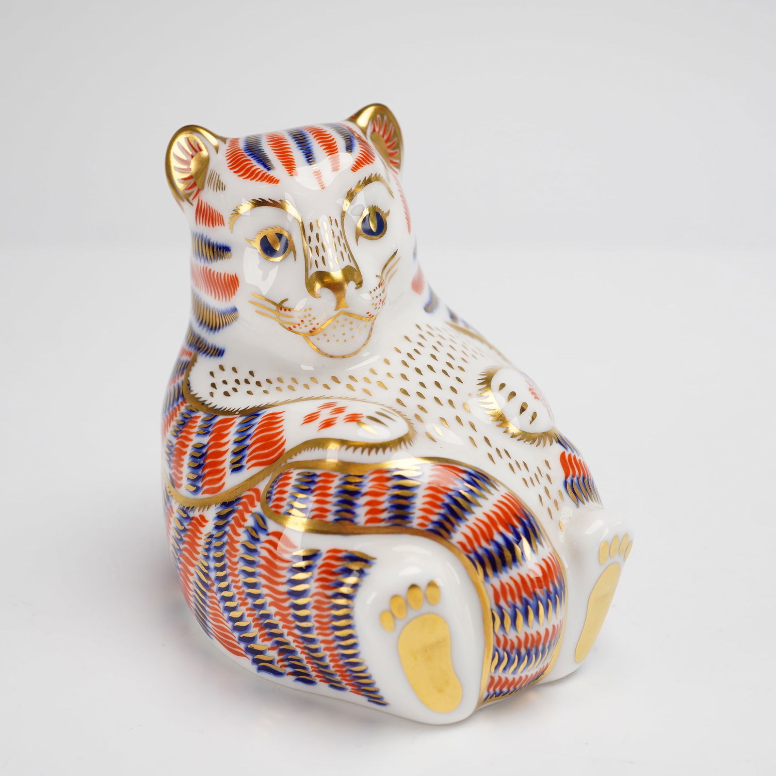 Royal Crown Derby Bone China Paperweight Tiger Cub: A Royal Crown Derby bone china paperweight depicting a seated tiger cub. The figurine is decorated in a stylized Imari pattern featuring orange and cobalt blue stripes with 22kt gold-gilt accents