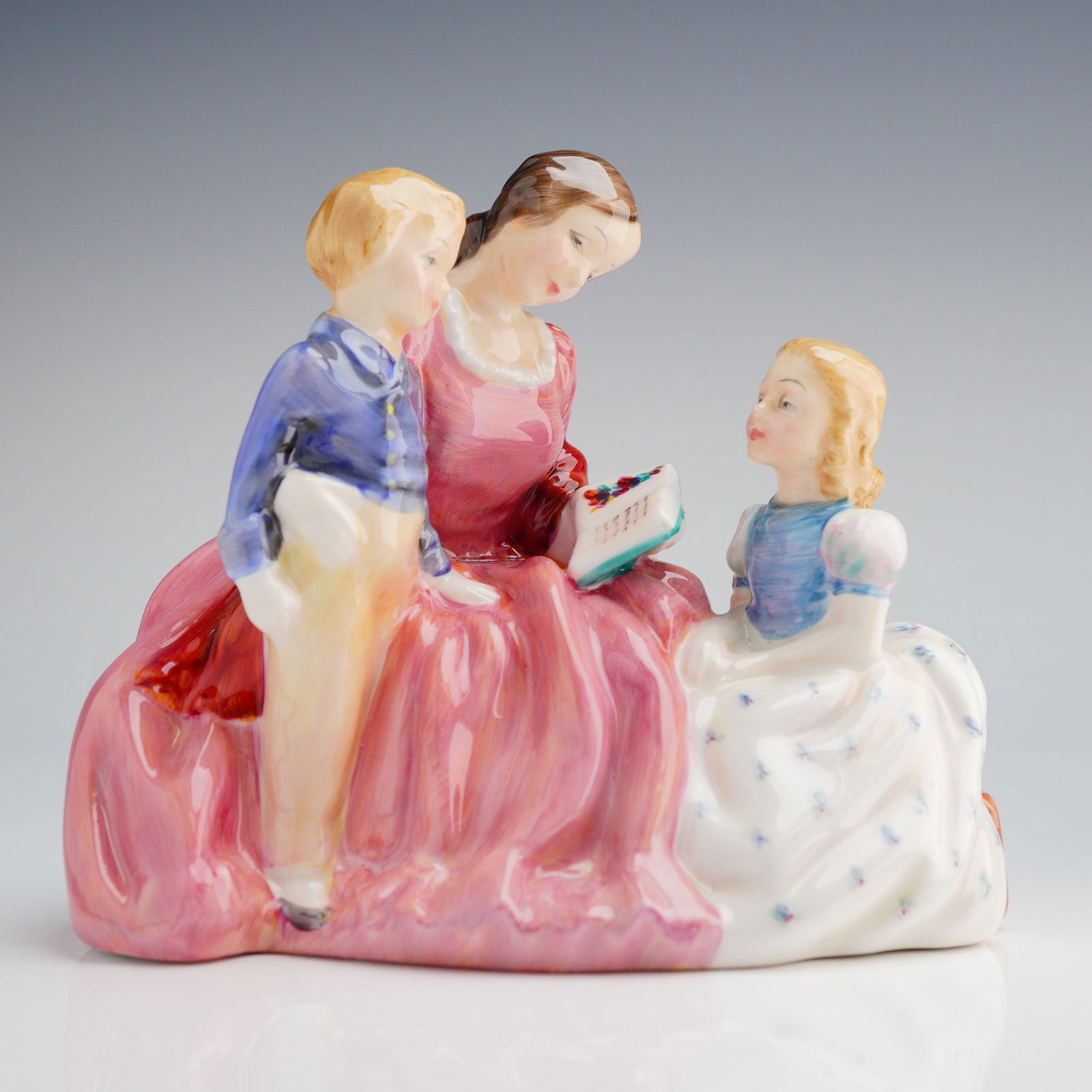 Royal Doulton Figurine, Bedtime Story HN2059: The piece depicts an adult woman seated, reading from a small book or sheet music to a young boy and a young girl seated beside her. The figures are highly glazed and hand-painted, featuring brightly