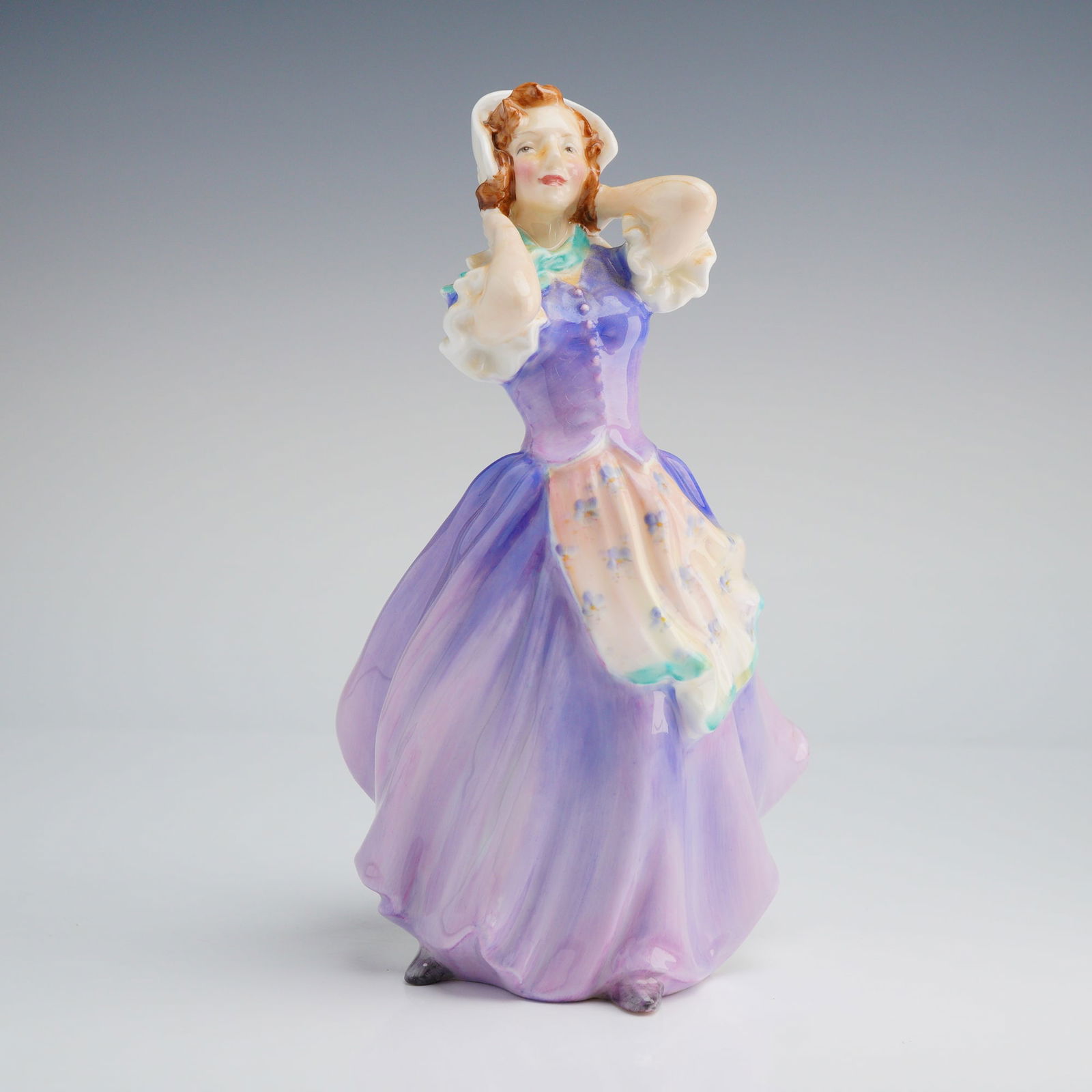 Royal Doulton Figurine, Betsy HN2111: The figurine depicts a standing young woman in a brightly colored costume, featuring a deep violet bodice and a voluminous skirt transitioning into lilac and purple hues, accented by a floral-patterne