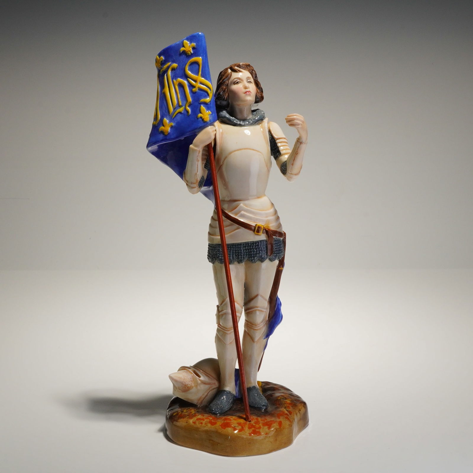 Royal Doulton Joan of Arc Porcelain Figurine HN3681: A glazed porcelain figurine manufactured by Royal Doulton, depicting Joan of Arc. The figure is shown standing in plate armor and chain mail, holding a long staff and a flowing cobalt blue banner deco