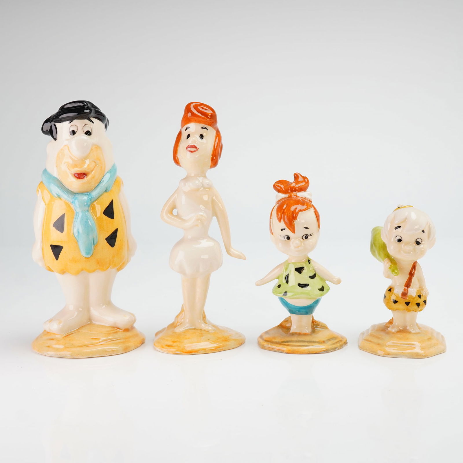 Set of 4 Beswick Porcelain Figurines, The Flinstone Family and Bamm-Bamm: The collection includes Fred Flintstone, Wilma Flintstone, Pebbles Flintstone, and Bamm-Bamm Rubble. Each figure is composed of glazed ceramic with a multi-colored finish and stands on an integral cir