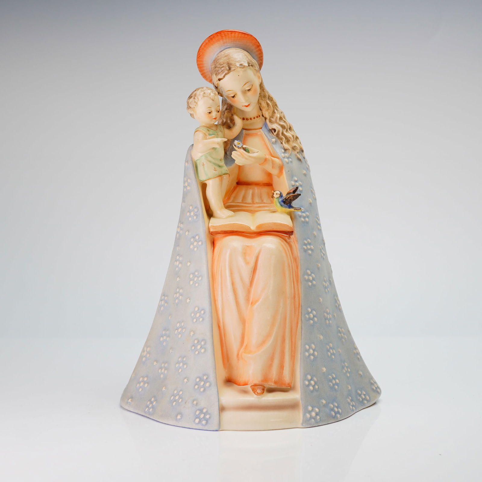 Goebel Hummel Figurine, Flower Madonna: The piece depicts the Virgin Mary seated with the Christ child and a bird perched upon an open book. The hand-painted figure features Mary in a light blue cloak decorated with white floral patterns ov