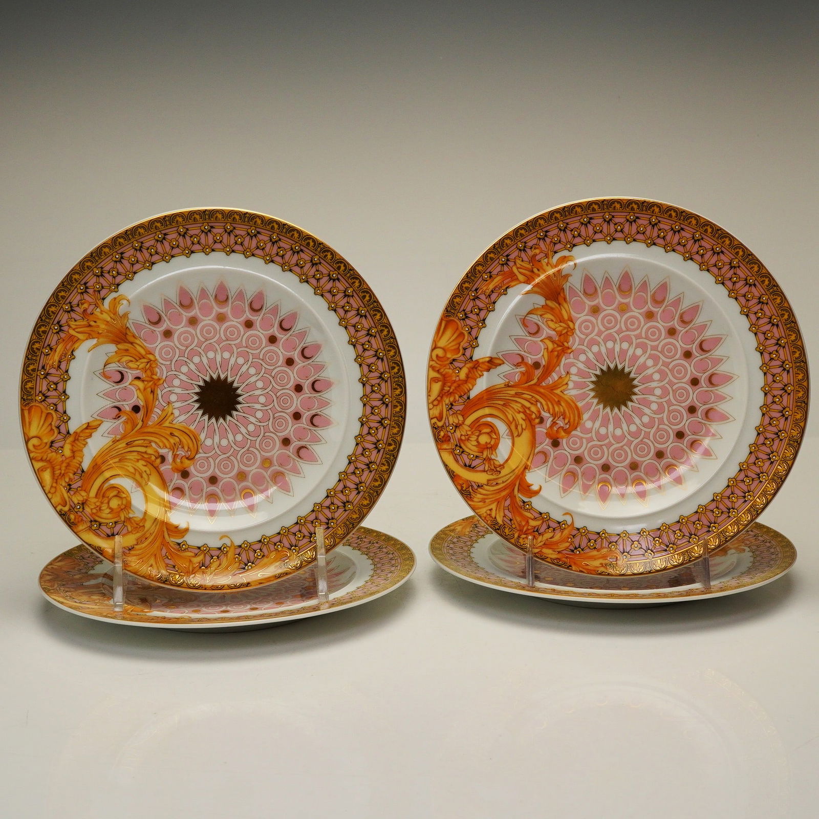Set of 4 Versace Rosenthal Porcelain Side Plates, Les Reves Byzantins: Each plate features a neo-baroque design consisting of gold-toned acanthus leaf scrolls layered over a central pink and white geometric sunburst motif, framed by a patterned pink and gold border. The