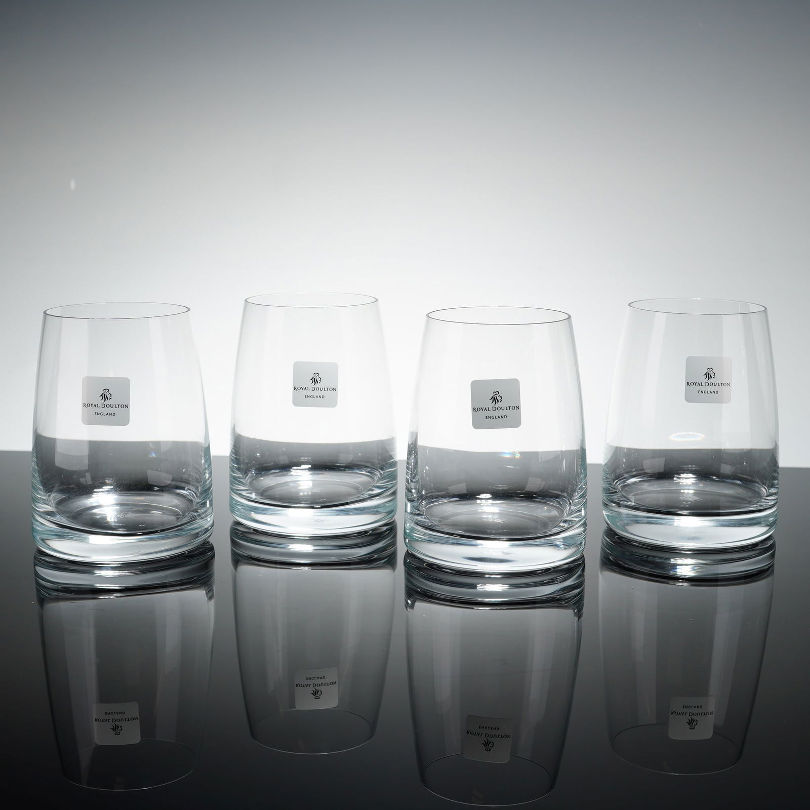 Set of 4 Royal Doulton Crystal Tumbler Glasses: Glasses feature a minimalist, slightly tapered cylindrical design. Each glass is constructed with a thick, weighted base. The items are marked with original silver-toned square labels displaying the m