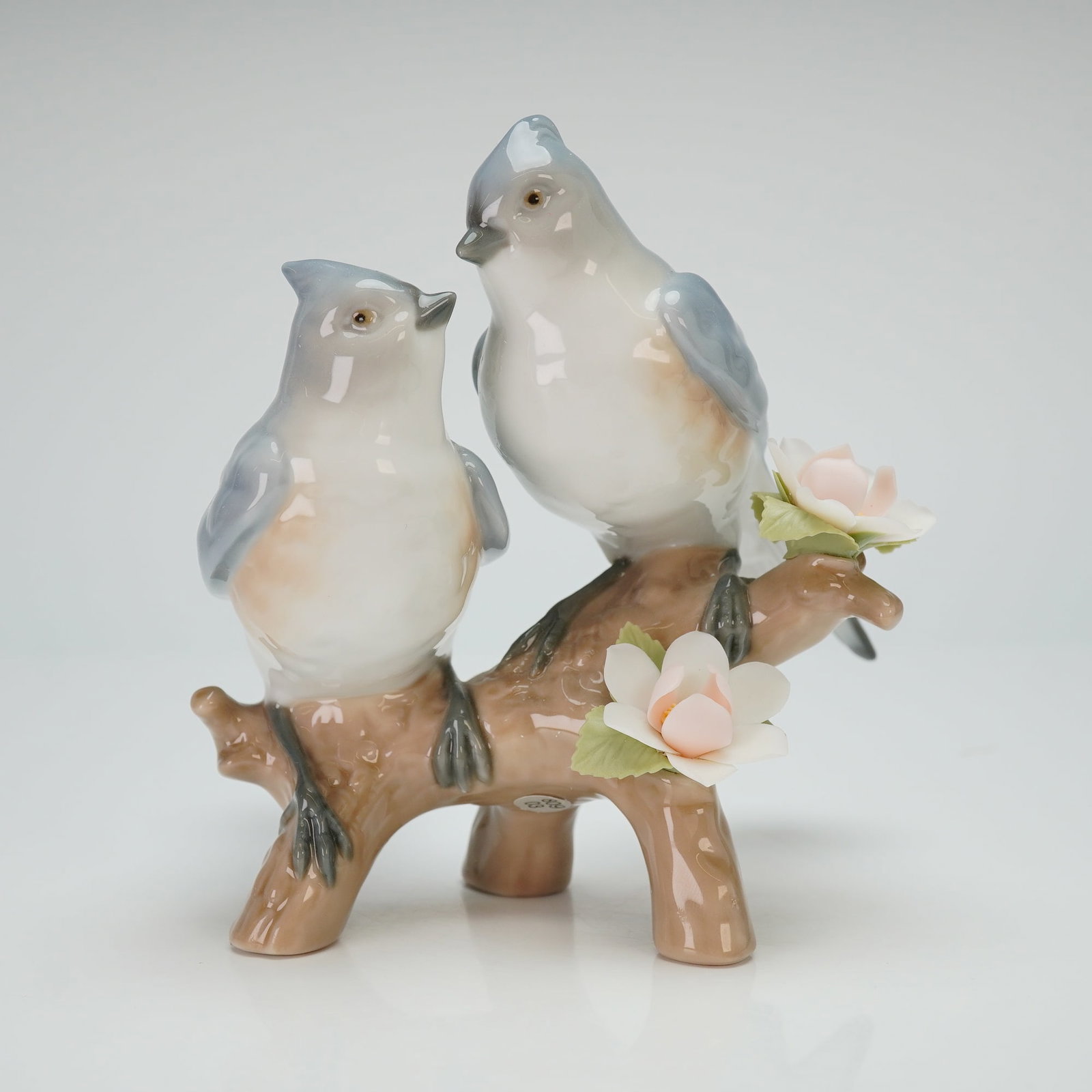 Lladro Porcelain Figurine Nature's Duet 01006917: This Lladro depicts two songbirds with soft, blue-toned plumage perched on a stylized branch. The composition is accented by two delicate pale pink blossoms and green leaves. The piece is executed in