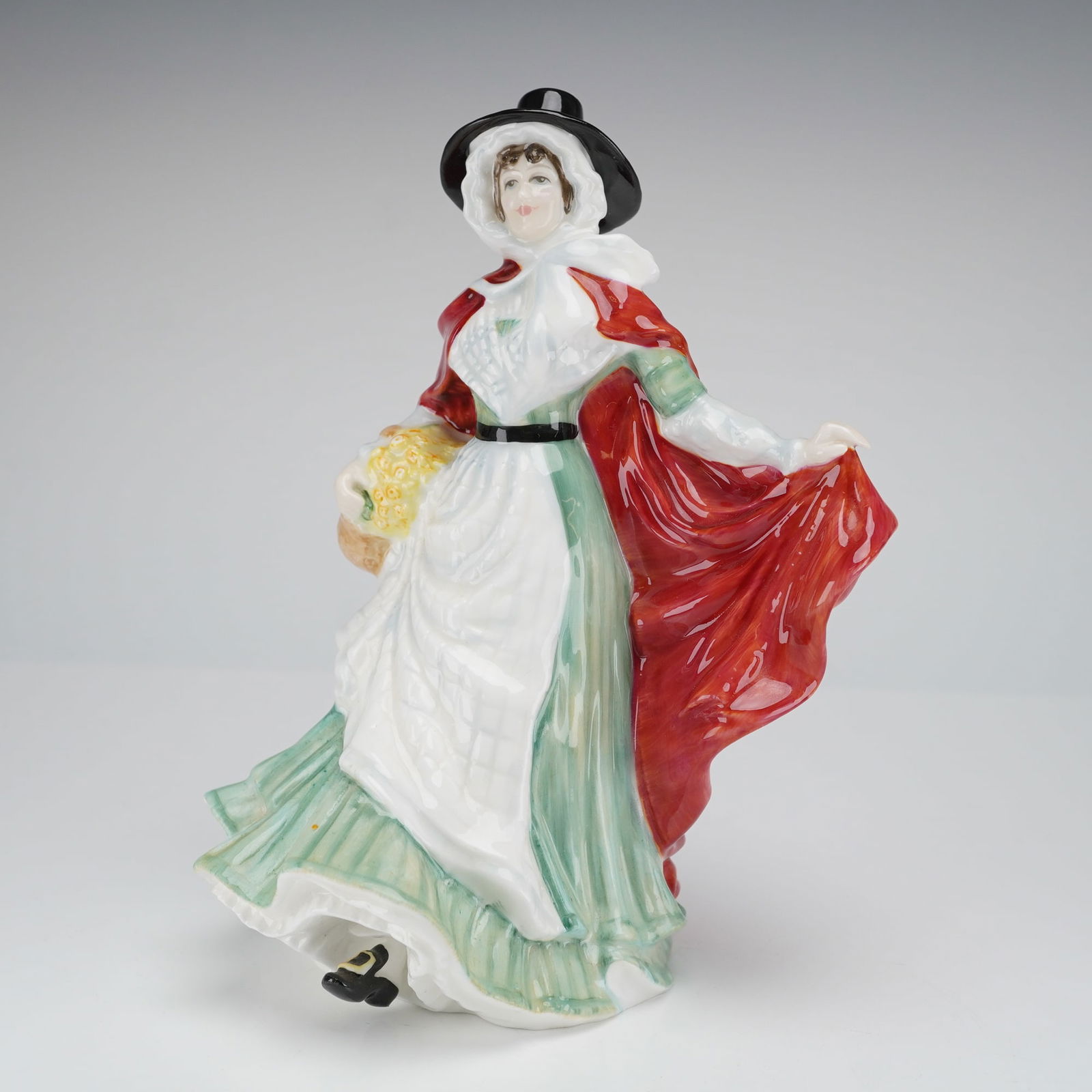 Royal Doulton Figurine, Ladies of the British Isles -Wales HN 3630: The figure is modeled as a traditional Welsh Lady, characterized by a high-crowned black hat, a white bonnet and apron, a mint green dress, and a flowing bright red cloak. She holds a basket containin