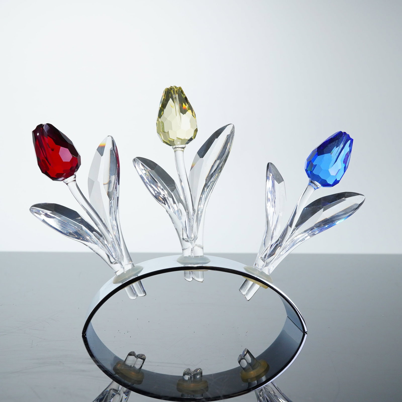 4pc Swarovski SCS Crystal Tulips and Display Stand: Swarovski Collector's Society (SCS) crystal tulips and matching display stand. The set includes faceted crystal tulips in shades of red, yellow, and blue, each designed with a multi-faceted colored fl