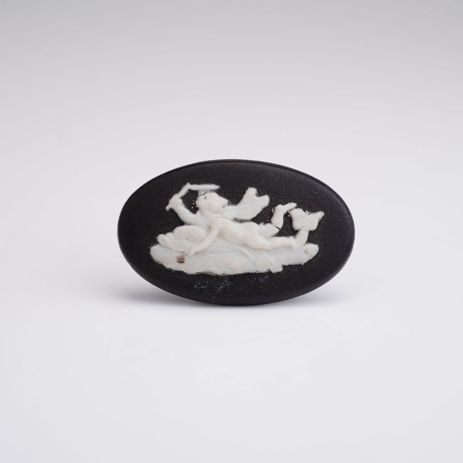 Wedgwood Black and White Jasperware Plaque of Cupid Riding on a Dolphin: A tiny oval plaque depicting Cupid riding a dolphin, a traditional Greco-Roman motif representing the swift and often turbulent passage of love and the soul across the sea of life. The reverse side is