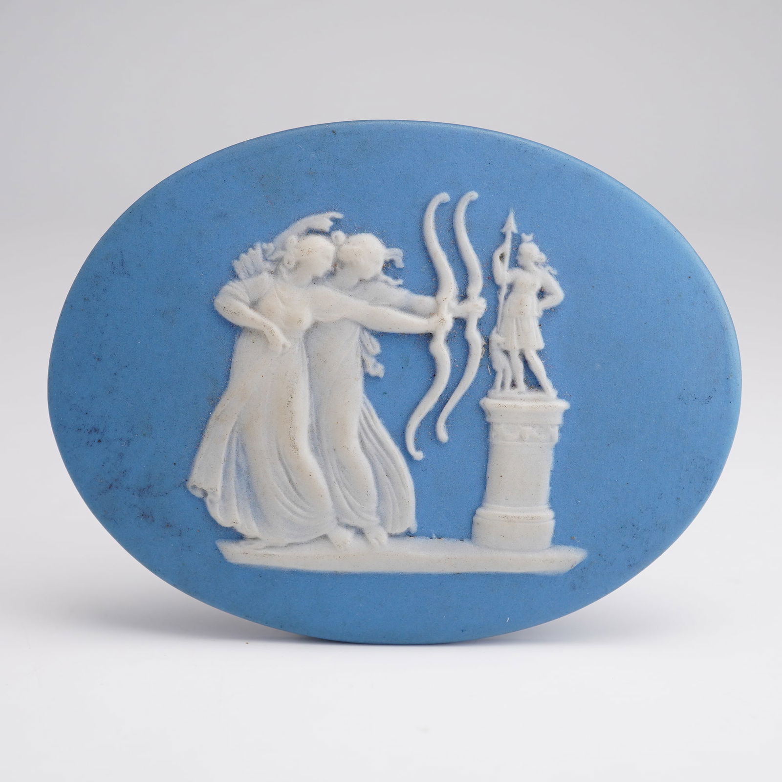 Wedgwood Blue and White Jasperware Plaque of the Archers: Small oval blue and white Jasperware plaque depicting two female archers posed to the left of a pedestal holding a statuette of the Greek goddess Artemis. The reverse side bears the "Wedgwood" impress