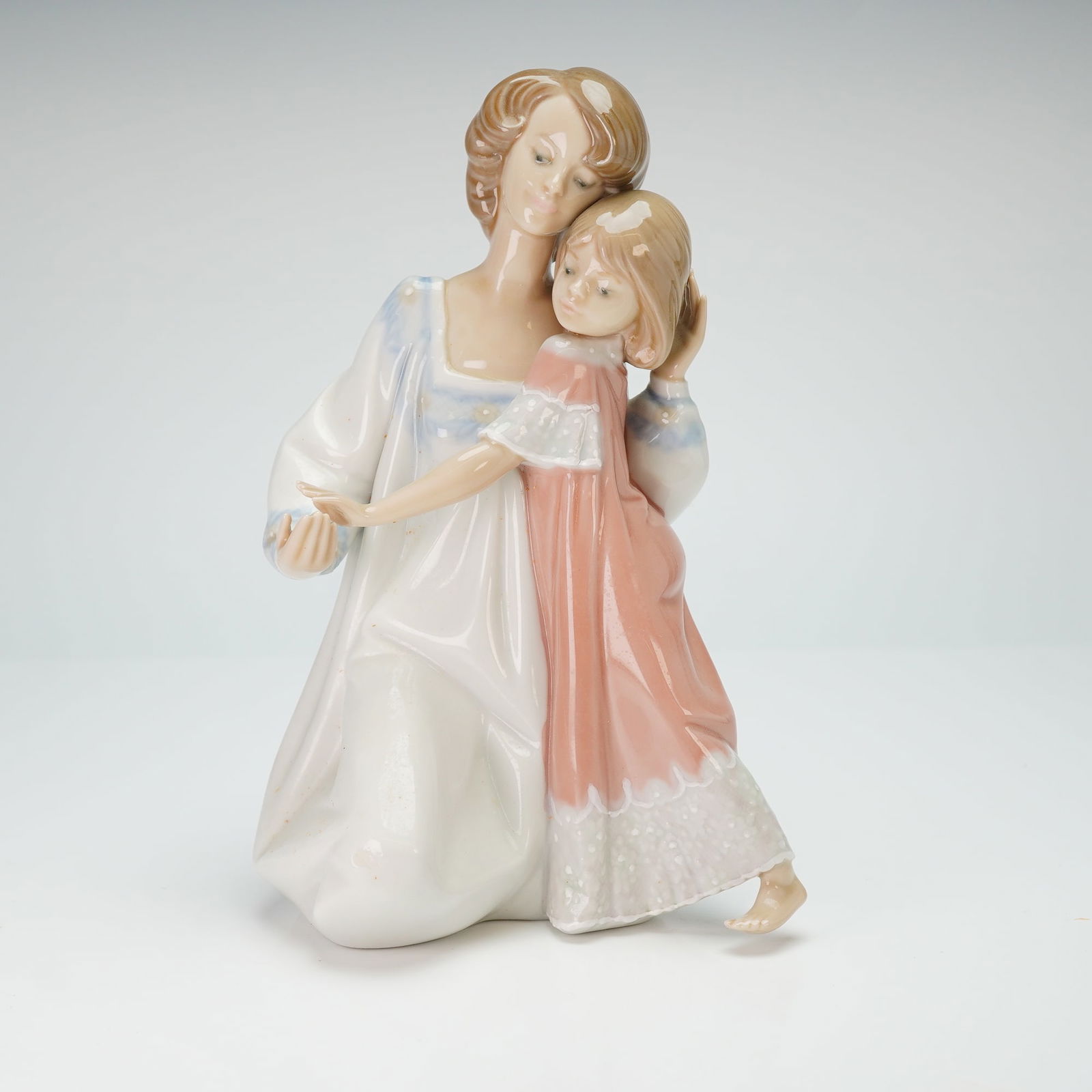 Lladro Porcelain Figurine Good Night 01005449: The piece depicts a mother kneeling while embracing a young child. The mother wears a long white gown with pale blue floral detailing at the neckline and cuffs, while the child is dressed in a soft pi