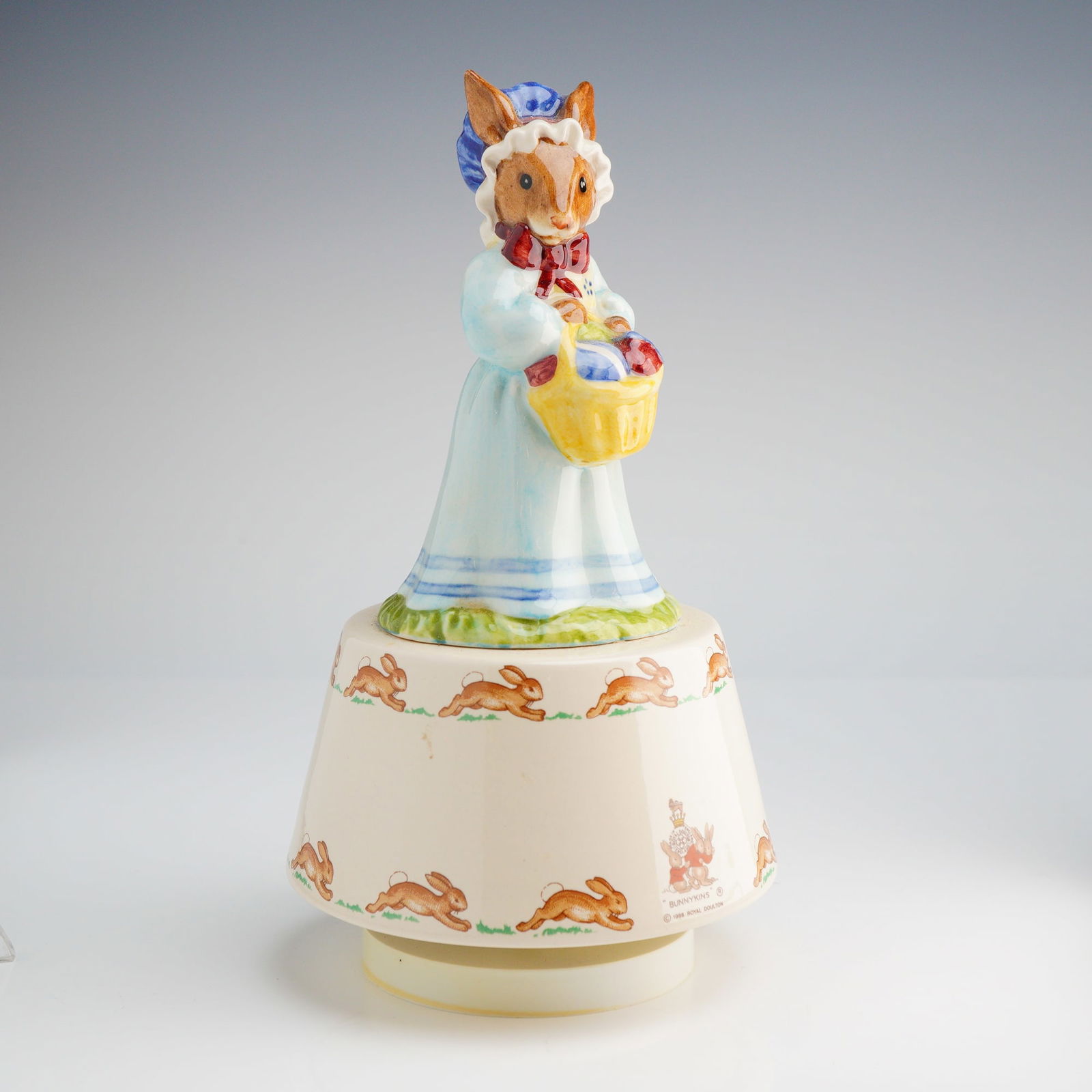 Royal Doulton Bunnykins Music Box, Easter Parade DB39: A Royal Doulton Bunnykins figural music box titled Mrs. Bunnykins at the Easter Parade, model number DB39. The ceramic piece features a hand-painted rabbit figurine dressed in a light blue gown and a