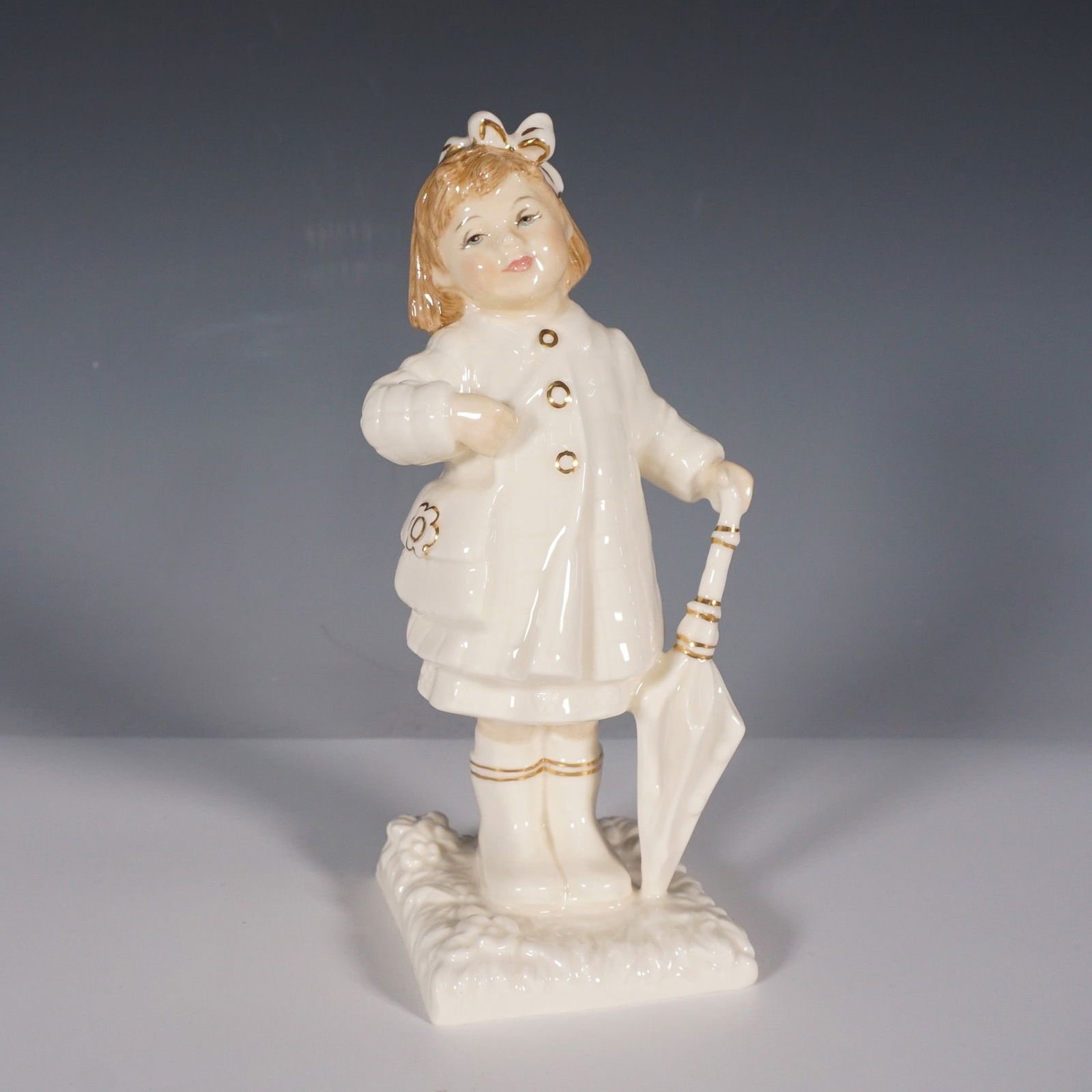 Royal Doulton Ivory and Gold Figurine, After The Rain HN4226: This charming figurine is part of the exclusive Ivory and Gold collection, showcasing a young girl standing in the aftermath of a rain shower. She is elegantly dressed in a detailed raincoat, boots, a