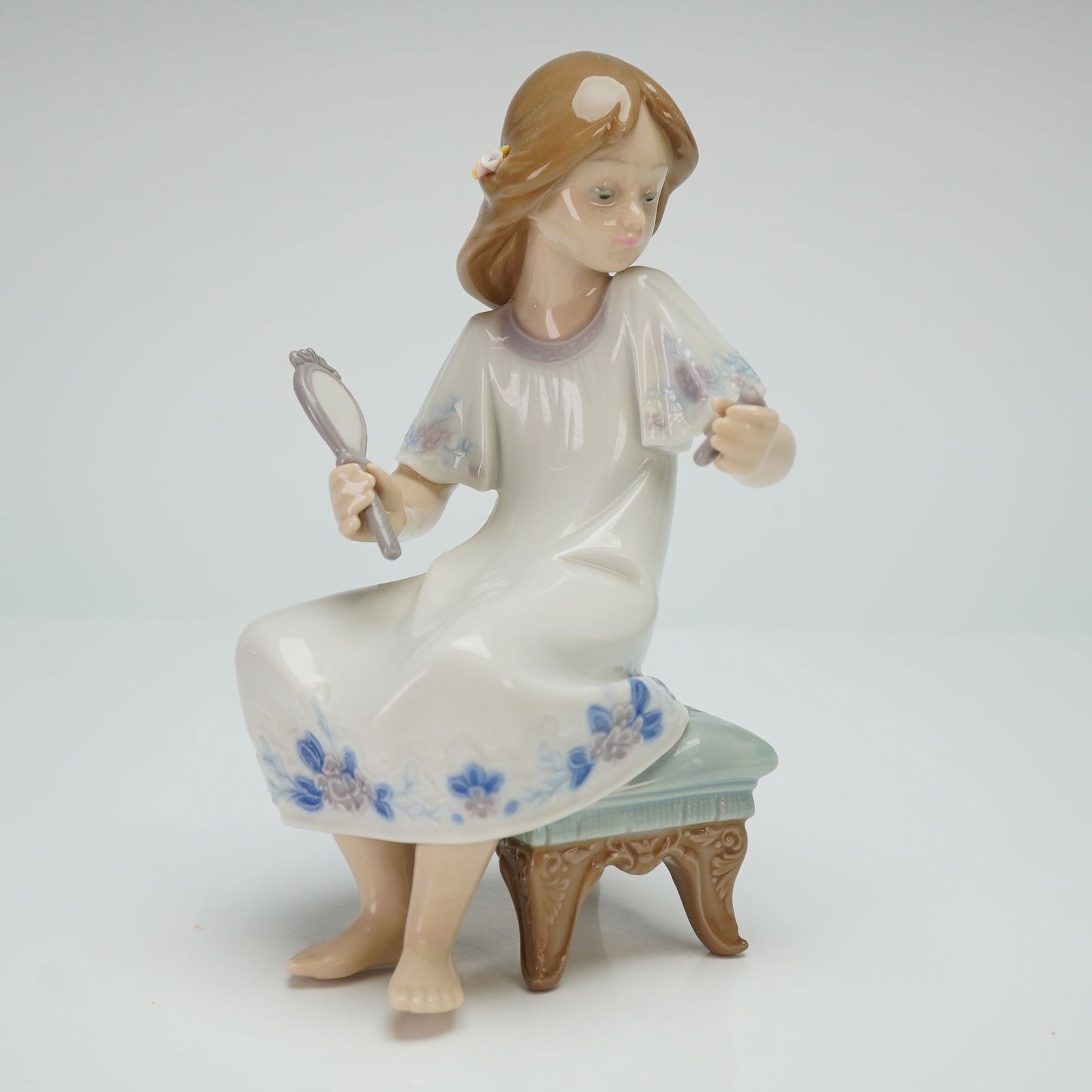 Lladro Porcelain Figurine I Feel Pretty 01005678: The figure depicts a young girl with long brown hair seated on an ornate light-blue cushioned footstool. She is dressed in a flowing white gown featuring blue and lavender floral motifs along the hem