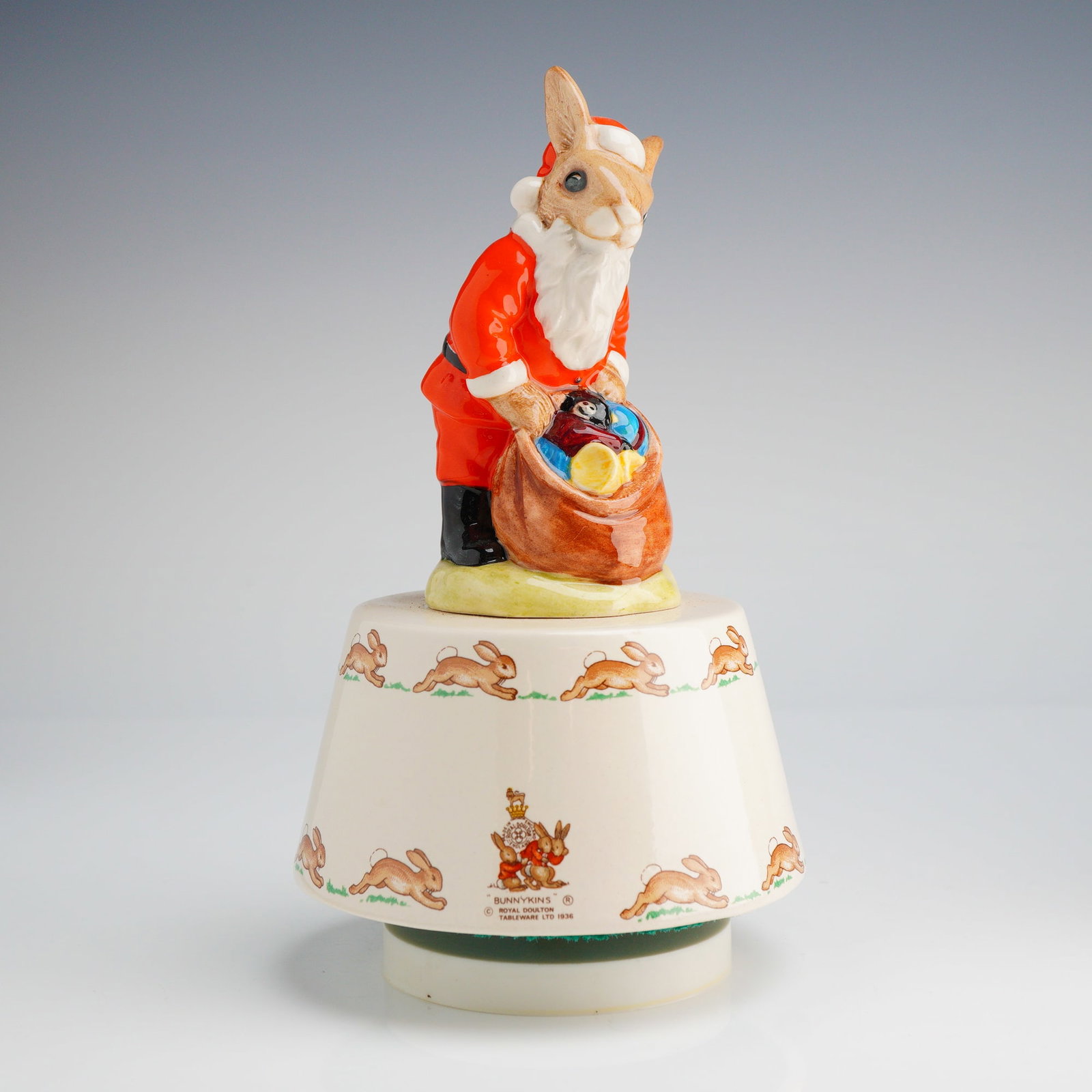 Royal Doulton Bunnykins Music Box, White Christmas DB34: A Royal Doulton Bunnykins musical figurine titled Santa Claus, model DB34. This glazed ceramic item depicts a rabbit in a Santa Claus costume holding a sack of toys, mounted on a domed cream-colored b