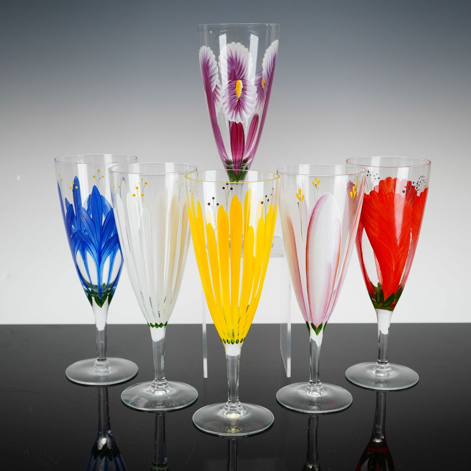 Set of Six Hand-Painted Glass Spring Flowers Wine Glasses: Each vessel is constructed of clear glass with a tapered, conical bowl supported by a slender stem and a flat circular base. The exterior of each bowl is decorated with hand-applied enamel depicting s