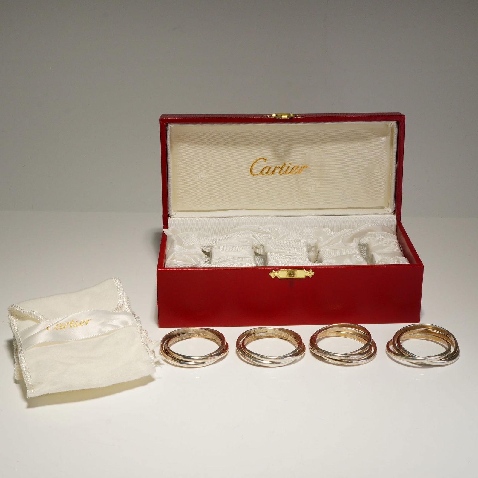 Set of Four Cartier Trinity Sterling Silver Napkin Rings: Napkin rings featuring three interlocked sterling silver circular bands. The design follows the classic Trinity motif. The set is presented in its original red rectangular Cartier box with a gold-tone