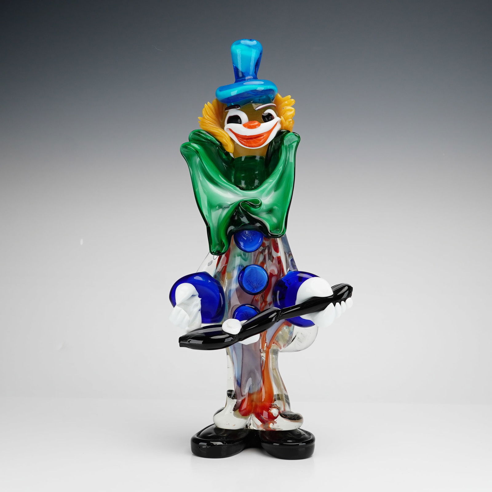 Murano Art Glass Clown Figurine: Clown figurine is depicted in a standing pose holding a black stringed guitar. The piece is hand blown from multi-colored glass and features a blue top hat, yellow textured hair, and a ruffled green b