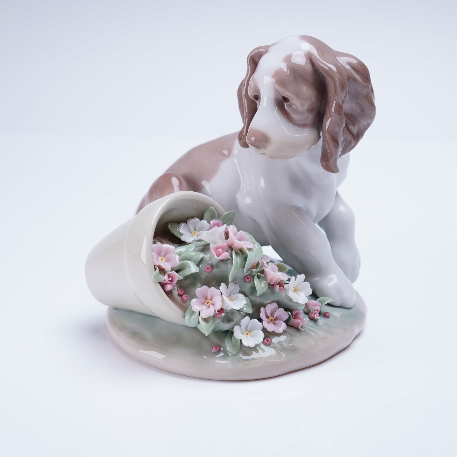 Lladro Figurine It Wasnt Me 1007672: The glossy glazed porcelain sculpture depicts a seated brown and white spaniel-type dog looking down intently at an overturned cream-colored flower pot. Highly detailed pink and white miniature flower