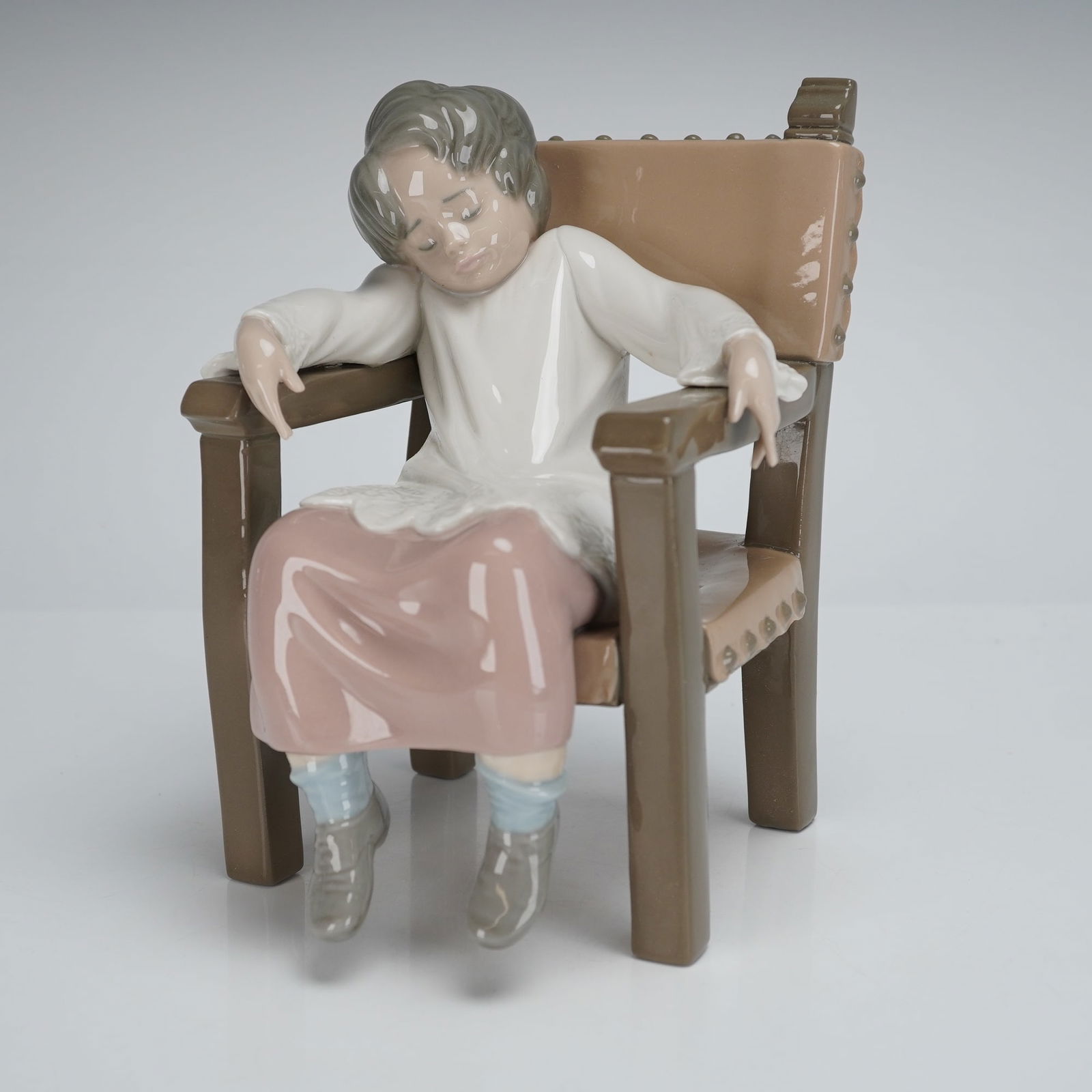 Lladro Porcelain Choir Boy Figurine 1005070: A single glazed Lladro Porcelain Figurine, titled "Choir Boy" model number 5070. The piece depicts a sleeping child slumped in a heavy, dark brown armchair. The chair features tan upholstery secured b