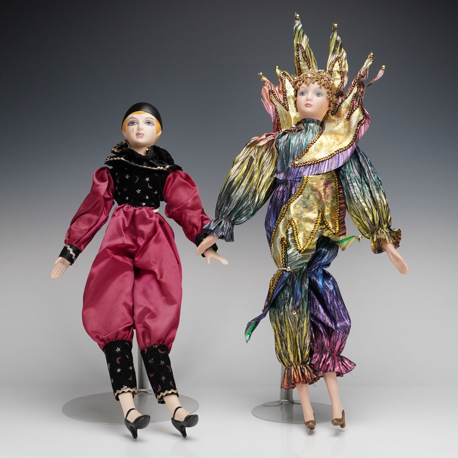A Pair of Porcelain Harlequin Collector Dolls: A pair of contemporary collector's dolls, featuring ceramic heads, hands, and lower limbs. The set includes a harlequin jester doll and a Mardi Gras-style doll. The jester doll is dressed in a theatri