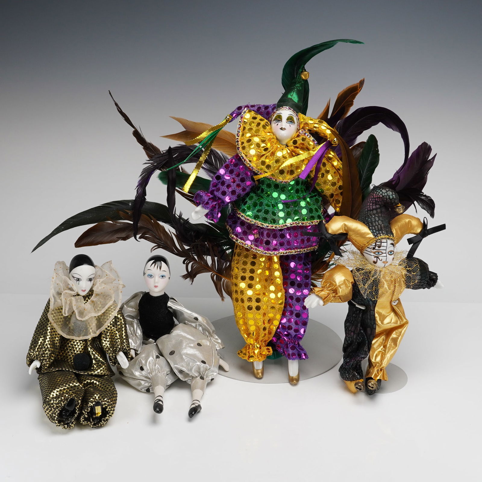 A Collection of Four Porcelain Harlequin Jester Dolls: A collection of four decorative porcelain harlequin dolls. The lot consists of four individual items, including two seated figures and two standing figures. Each doll features a painted porcelain head