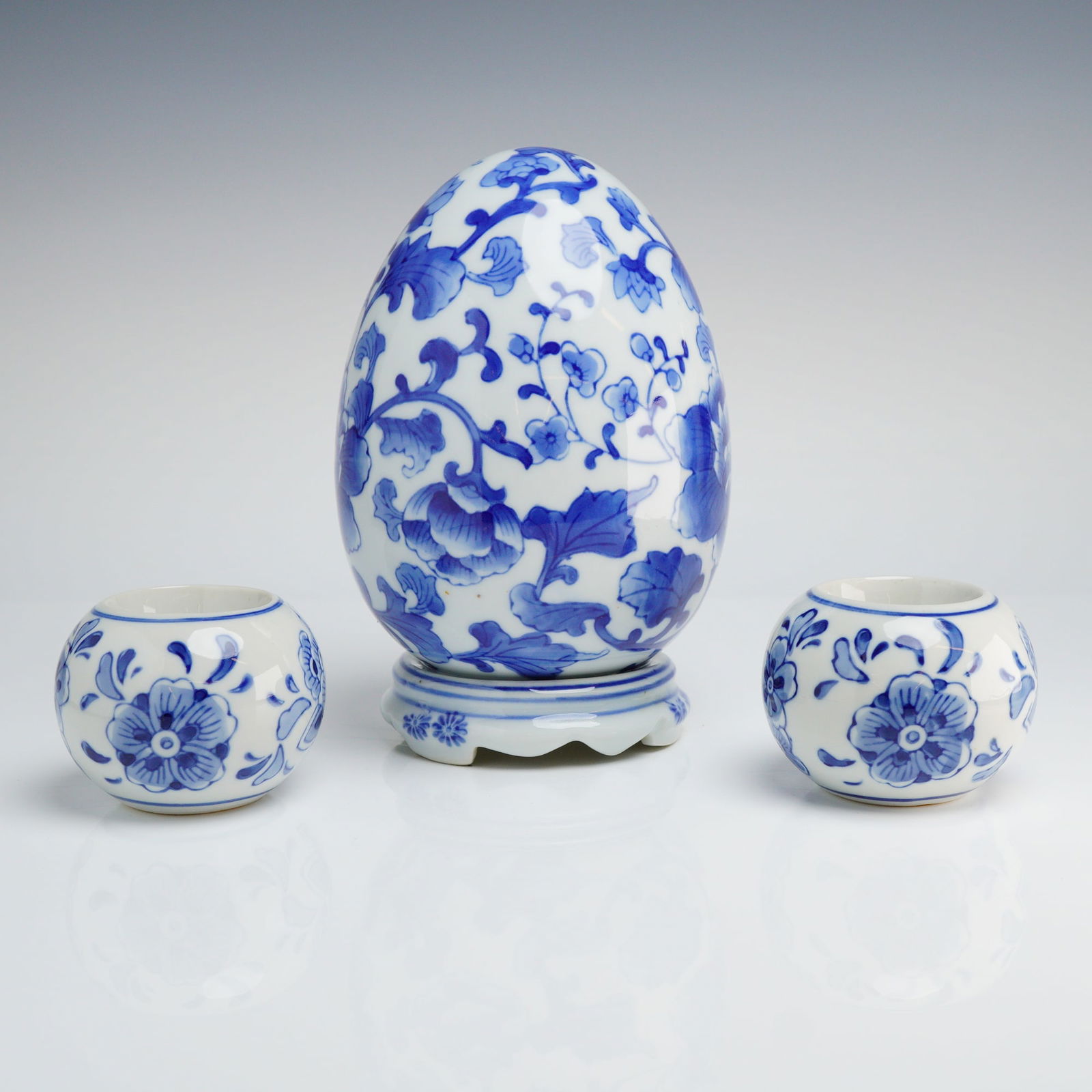 3pc Blue and White Chinoiserie Ceramic Egg and Candleholders: A collection of contemporary ceramic decorative objects executed in the blue and white Chinoiserie style. The grouping comprises one large egg-shaped footed ornamental piece, and a pair of small, sphe