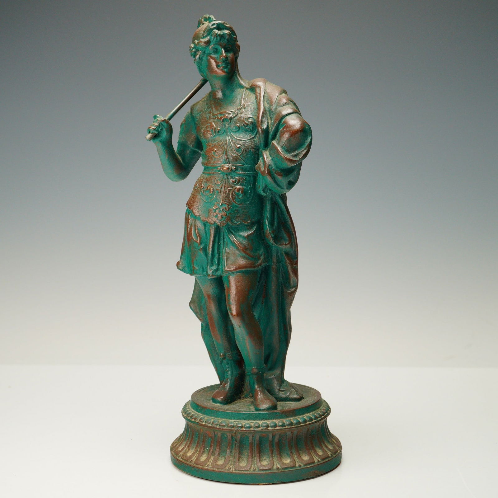 Patinated Metal Roman Soldier Sculpture: Cast metal sculpture with a rich green and bronze patina, featuring a classical figure in detailed armor and draped cloth, posed with a staff across the shoulder, standing atop a fluted round base