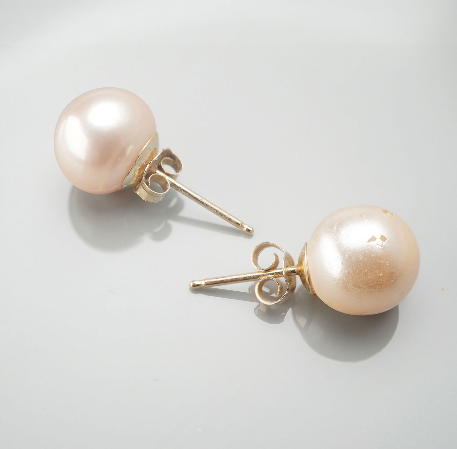 14K Gold Pearl Stud Earrings: Round light peach pearl studs mounted on 14K gold posts with matching 14K stamped butterfly backs. Each pearl set in a simple gold cup mounting. Posts and backs marked 14K. Total combined weight: 1.7