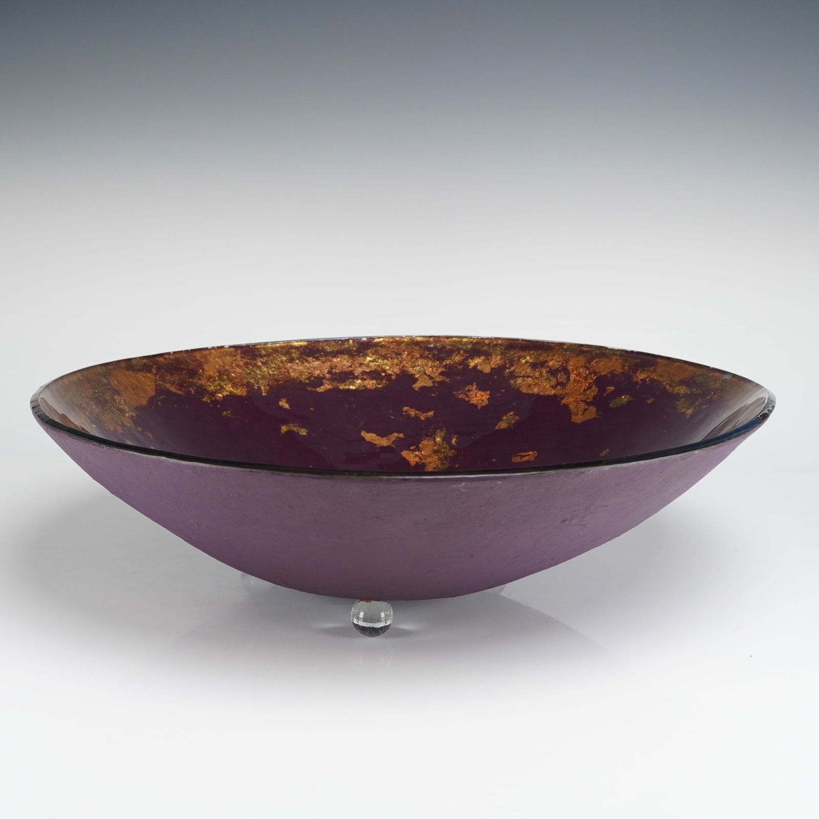 Tony Evans Fused Art Glass Bowl with Gold Leaf Inclusions: The piece is a contemporary fused art glass bowl by Tony Evans. The shallow, wide form features a deep plum-colored interior surface, glossy in finish, heavily decorated with fragmented applications o