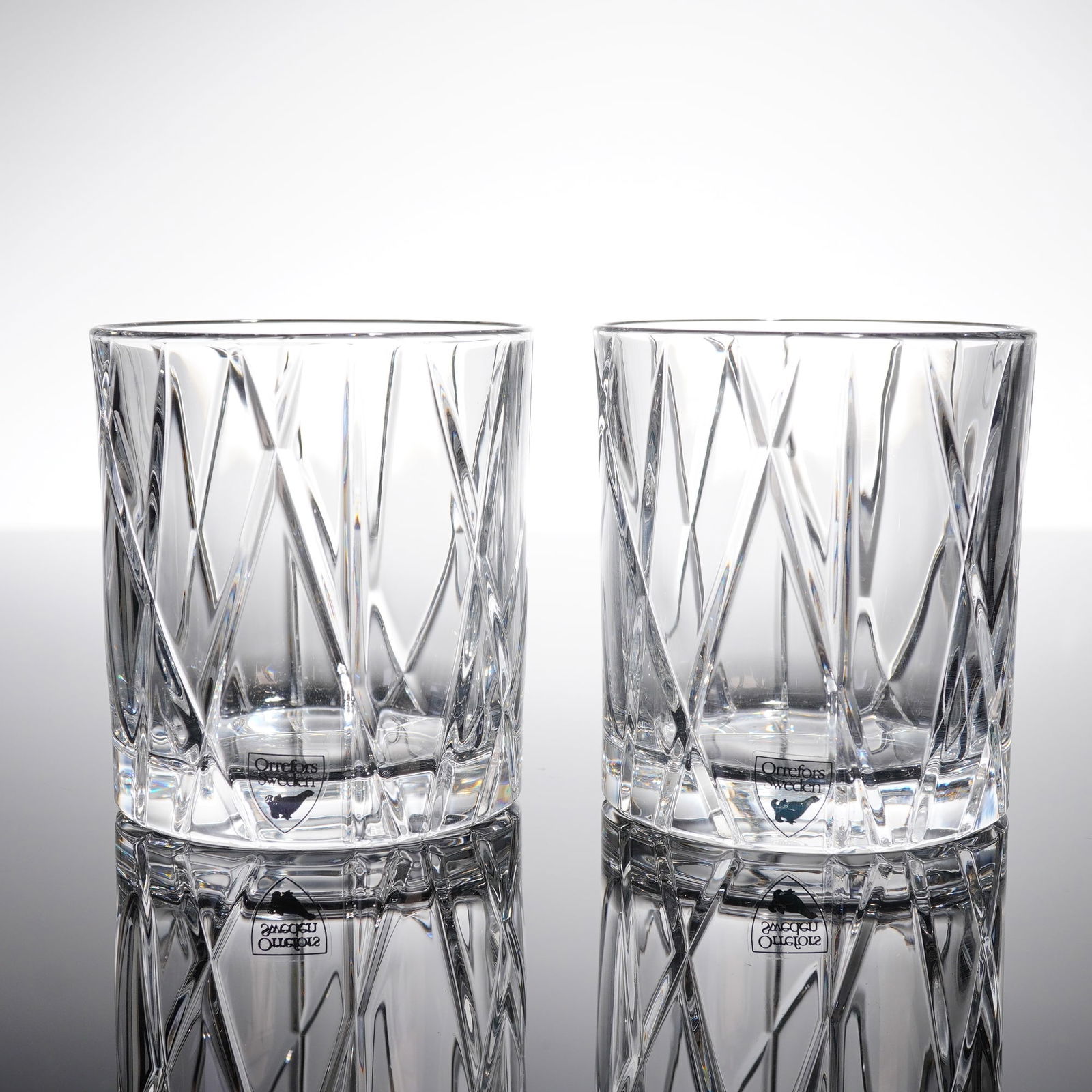 A Pair of Orrefors City Cut Crystal Double Old Fashioned Glasses: A pair of Orrefors City double old fashioned whiskey glasses crafted from clear cut crystal. Each tumbler features a cylindrical form with a thick, smooth base. The body is extensively decorated with