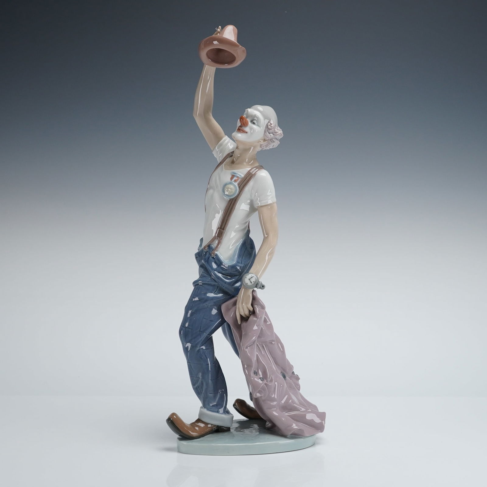 Lladro Hats Off to Fun Sculpture 1005765: This Lladro porcelain figurine is executed in a glossy glazed finish and depicts a contemporary clown figure standing, wearing a white t-shirt and blue patched overalls, with brown shoes. The clown
