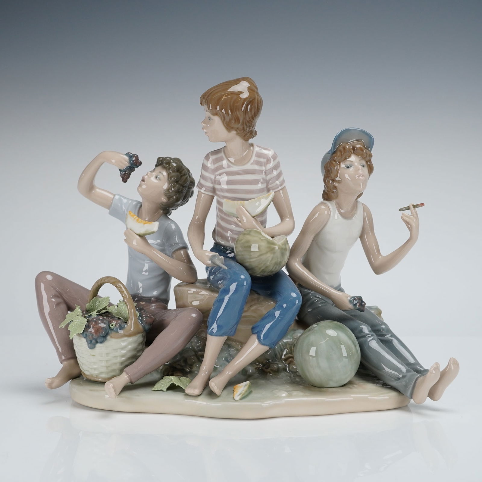Lladro Sweet Harvest Figurine 1005380: The Lladro porcelain figurine depicts three casually dressed boys seated upon a rocky outcrop, engaging with fruit. The figures are rendered in high-gloss polychrome porcelain. One boy is shown