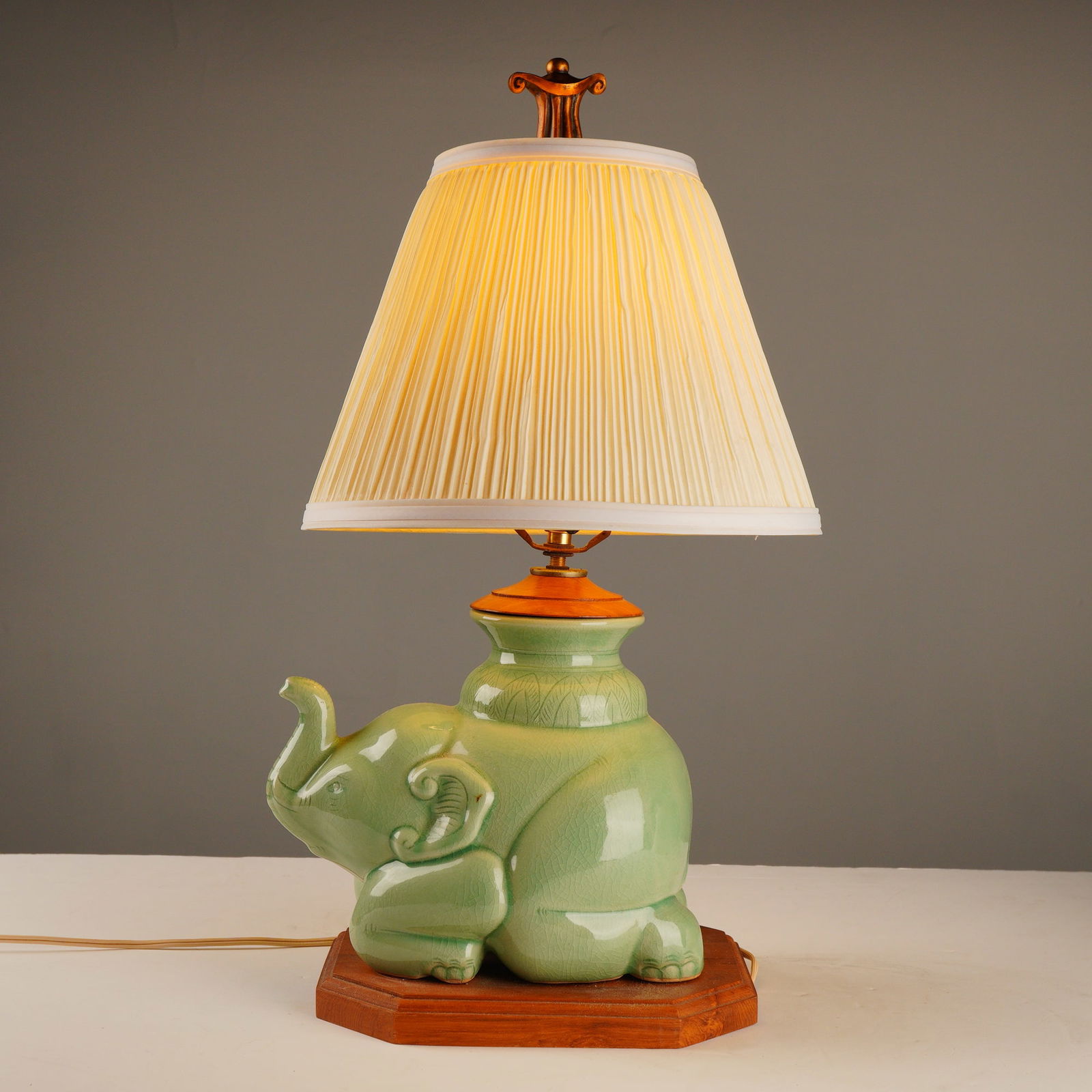 East Asian Celadon Ceramic Elephant Figural Table Lamp: A table lamp featuring a figural base sculpted in the form of a kneeling elephant. The ceramic body is finished with a pale celadon green crackle glaze, characteristic of East Asian ceramics. The elep