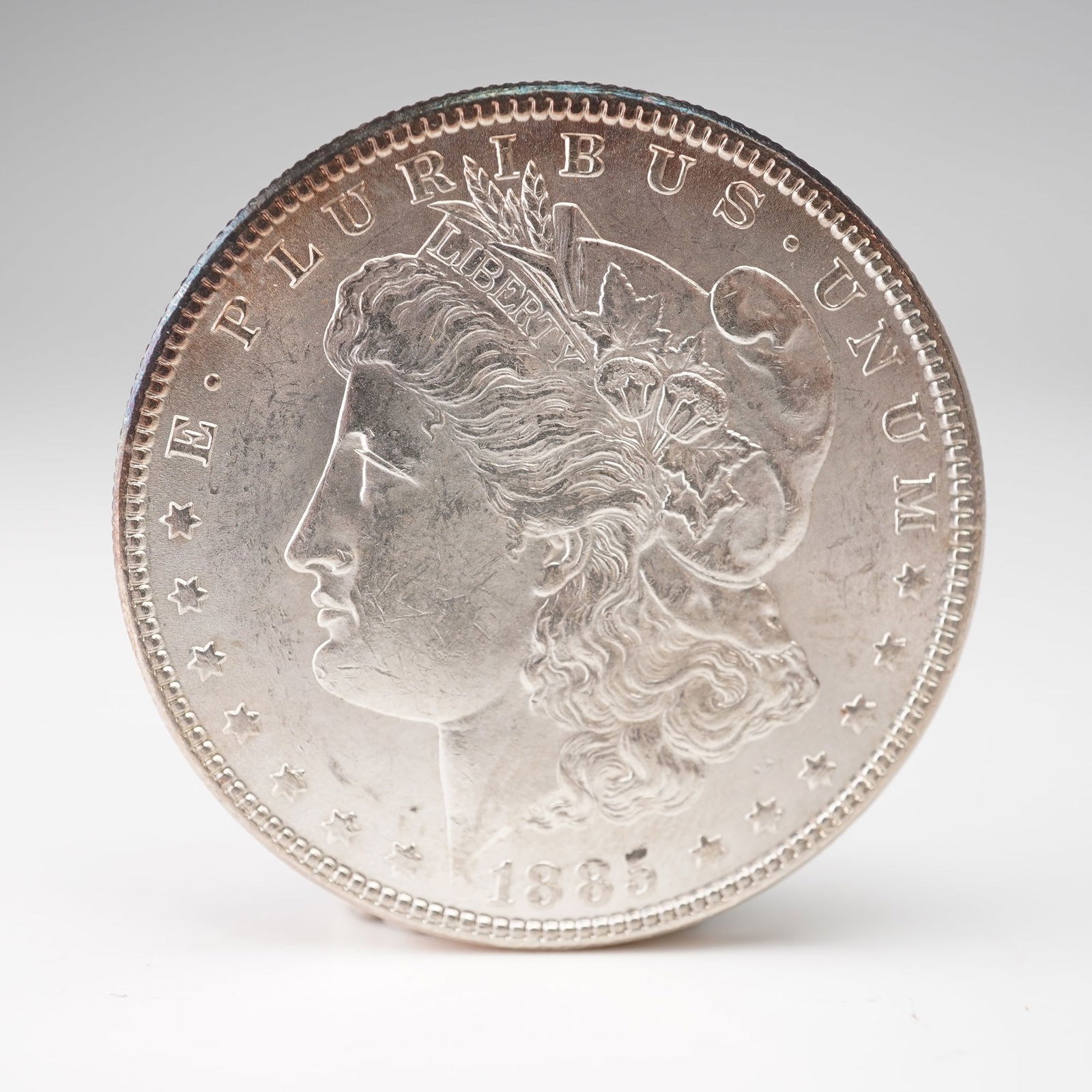 1885 Morgan Silver Dollar: An 1885 Morgan Silver Dollar, designed by George T. Morgan. The obverse features the elegant profile of Liberty, crowned with a Phrygian cap entwined with wheat and cotton, and is encircled with E PLU