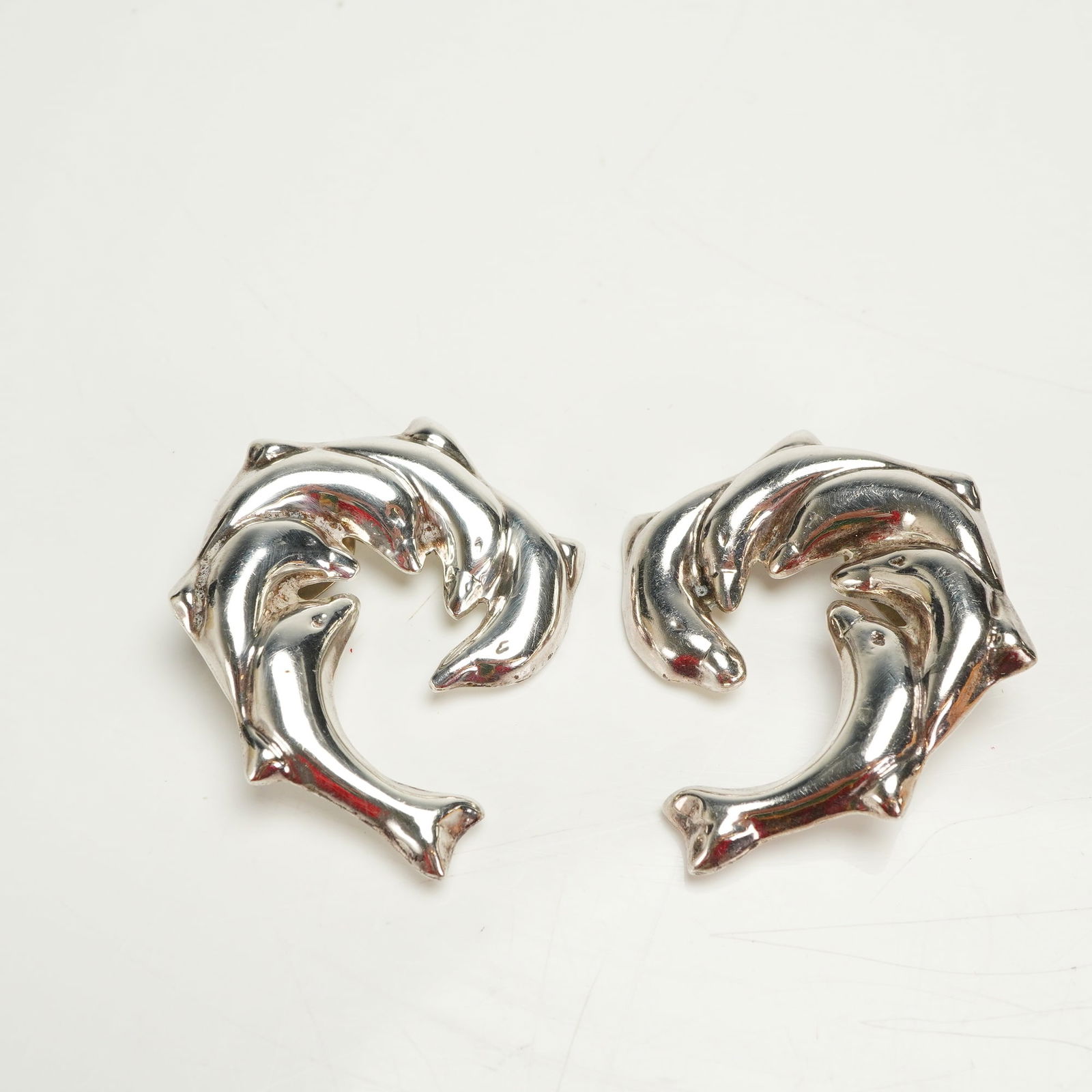 A Pair of Mexican Sterling Silver Modernist Dolphin Clip-On Earrings: A pair of sterling silver clip-on earrings crafted in Mexico. The design features a modernist, figural motif depicting two highly stylized dolphins or porpoises intertwined, forming a curved, open sha