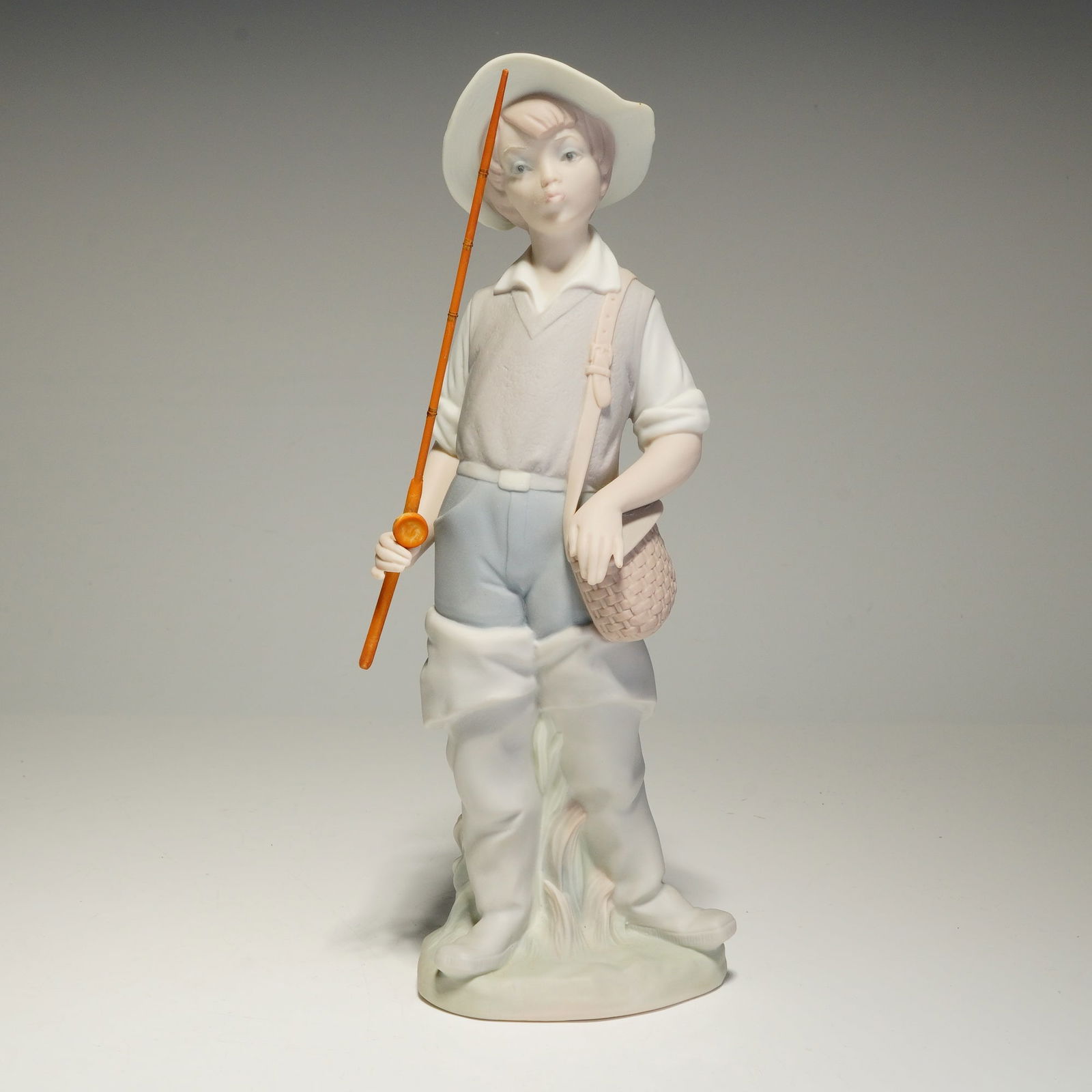 Lladro Matte Porcelain Figurine, Gone Fishing 01014809: A Lladro matte-finish porcelain figurine designed by sculptor Salvador Furio. The piece depicts a young boy dressed as a fisherman, standing in a composed pose. He is outfitted in a wide-brimmed hat,