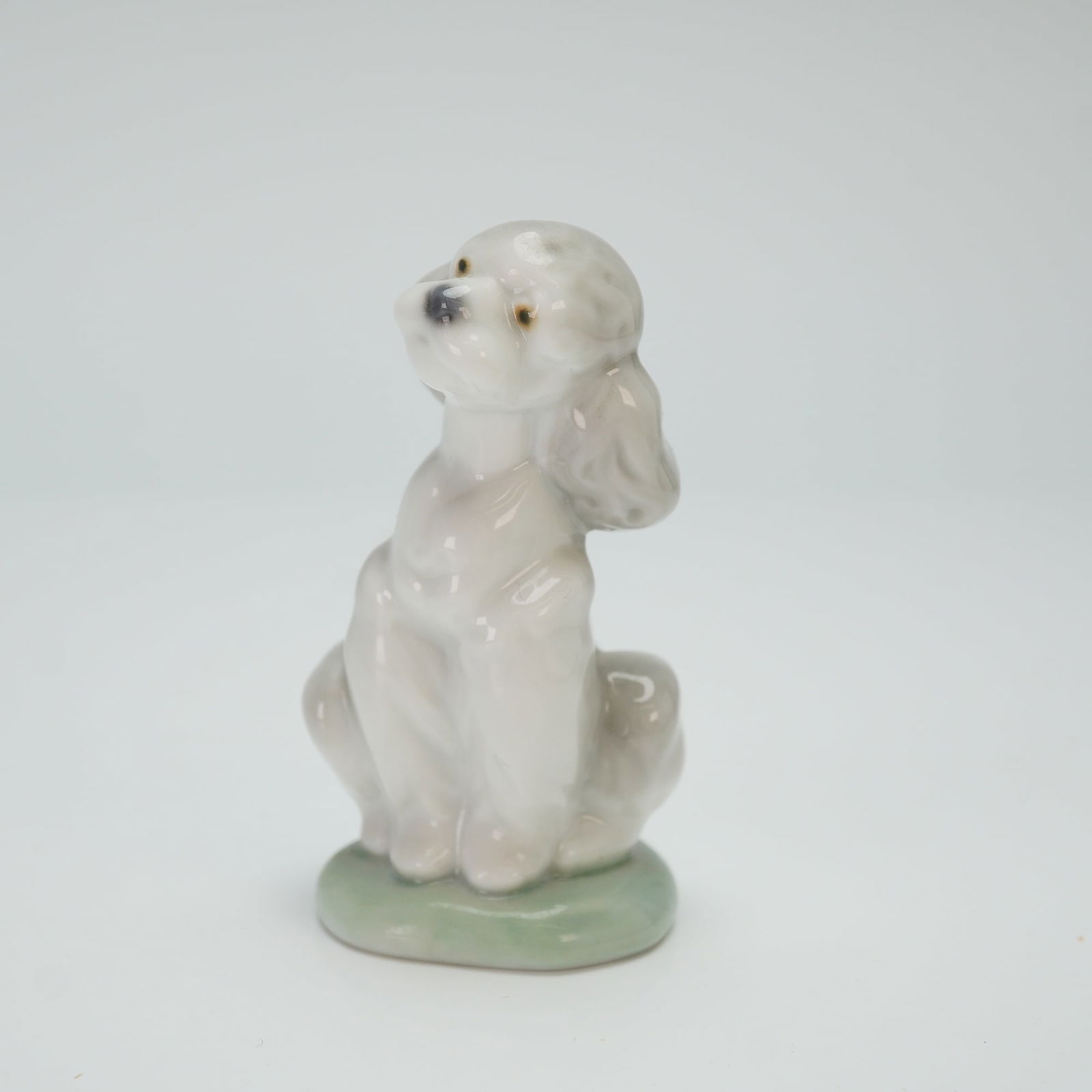 Lladro Porcelain Figurine A Friend for Life 01007685: This piece depicts a seated poodle looking upward on an integral pale green base. The figure is finished in a high-gloss glaze of white and pale gray tones. The underside features the blue circular Ll