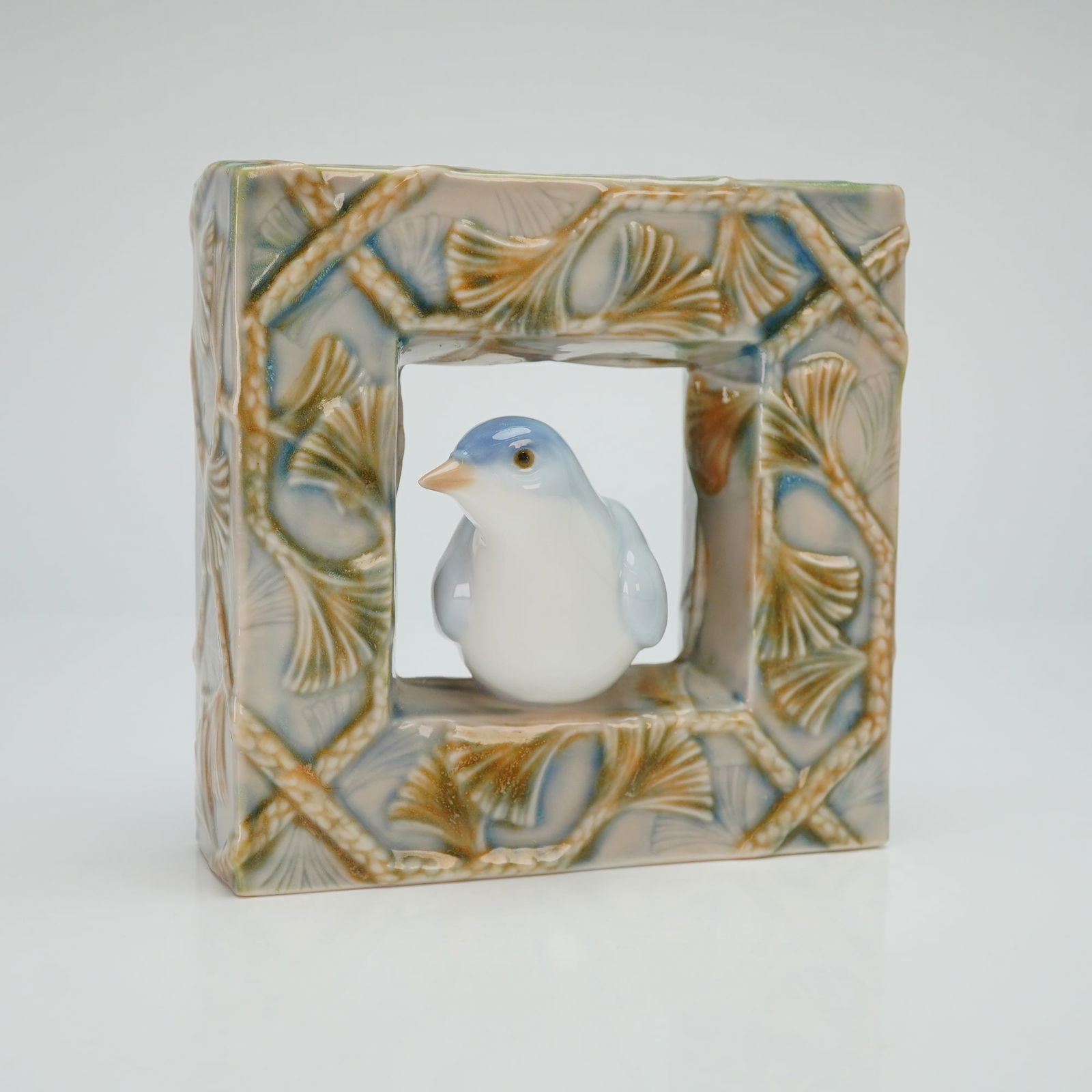 Lladro Porcelain Figurine Bird Natural Frames 01008067: This glazed porcelain figurine features a pale blue and white bird perched within a square, open-work frame. The frame is decorated with a relief ginkgo leaf and trellis pattern and is finished in a p