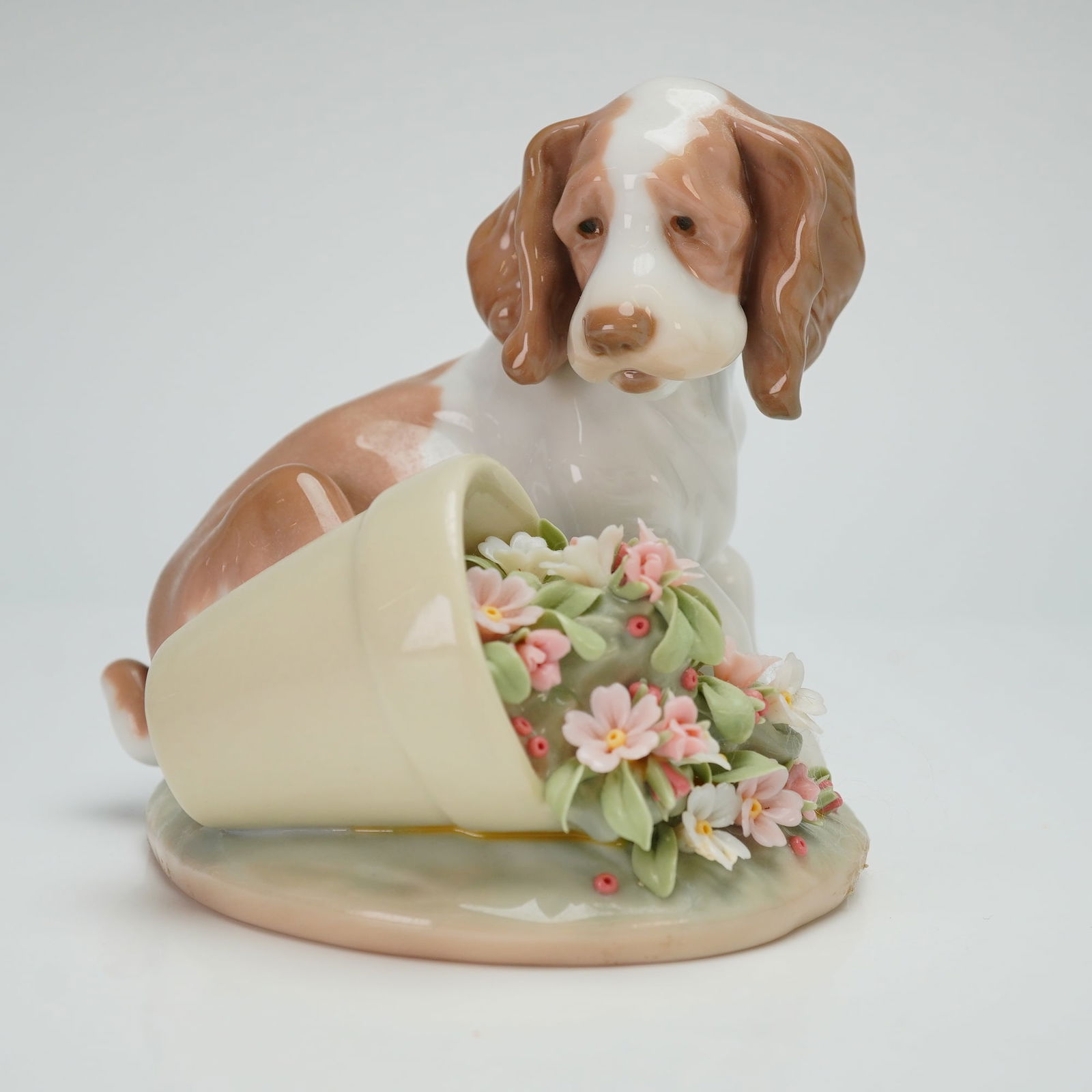 Lladro Porcelain Figurine It Wasn't Me 01007672: A Lladro issued as the 1998 Lladro Collectors Society membership piece. The glazed porcelain work depicts a tan and white spaniel puppy positioned on a circular base next to an overturned flowerpot. T