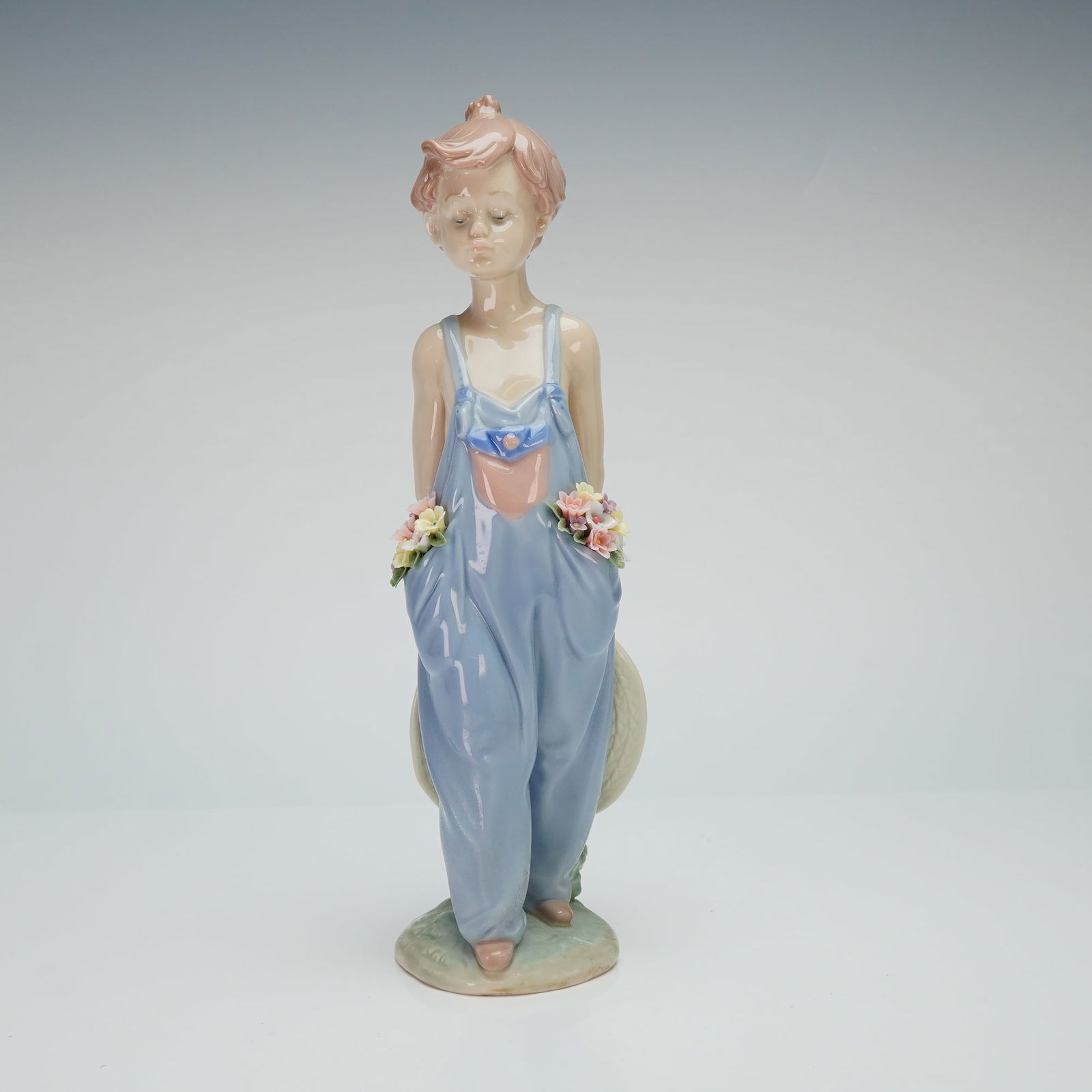 Lladro Porcelain Figurine Pocket Full of Wishes 01007650: A Lladro issued by the Lladro Collectors Society. The figure portrays a young boy wearing oversized blue overalls with pockets containing small, polychrome applied flowers. The boy is shown holding a