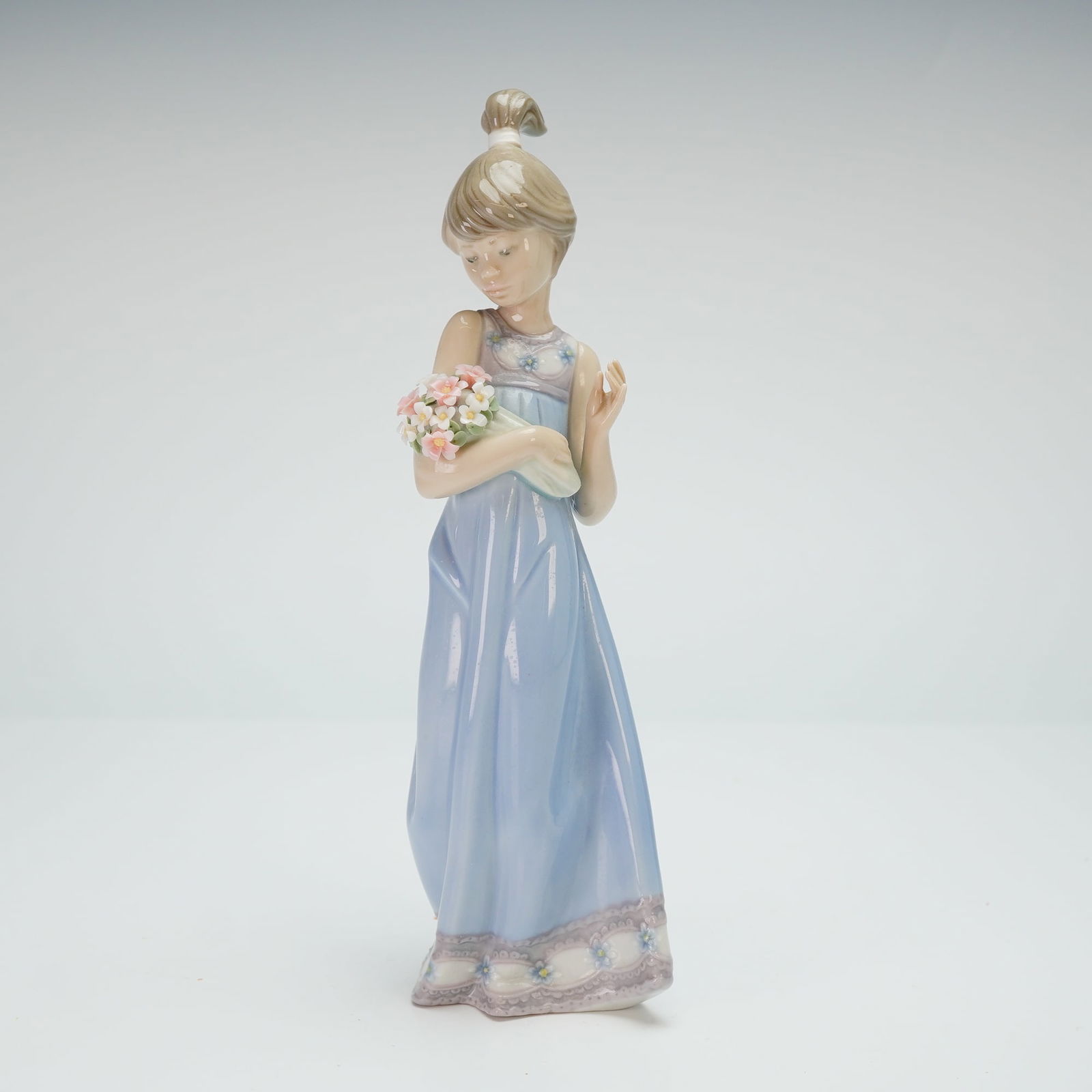 Lladro Porcelain Figurine Spring Token 01005604: The figure depicts a girl in a light blue sleeveless gown holding a bouquet of pink and white blossoms. The figure is styled with a high ponytail. The dress features a decorative floral border at the