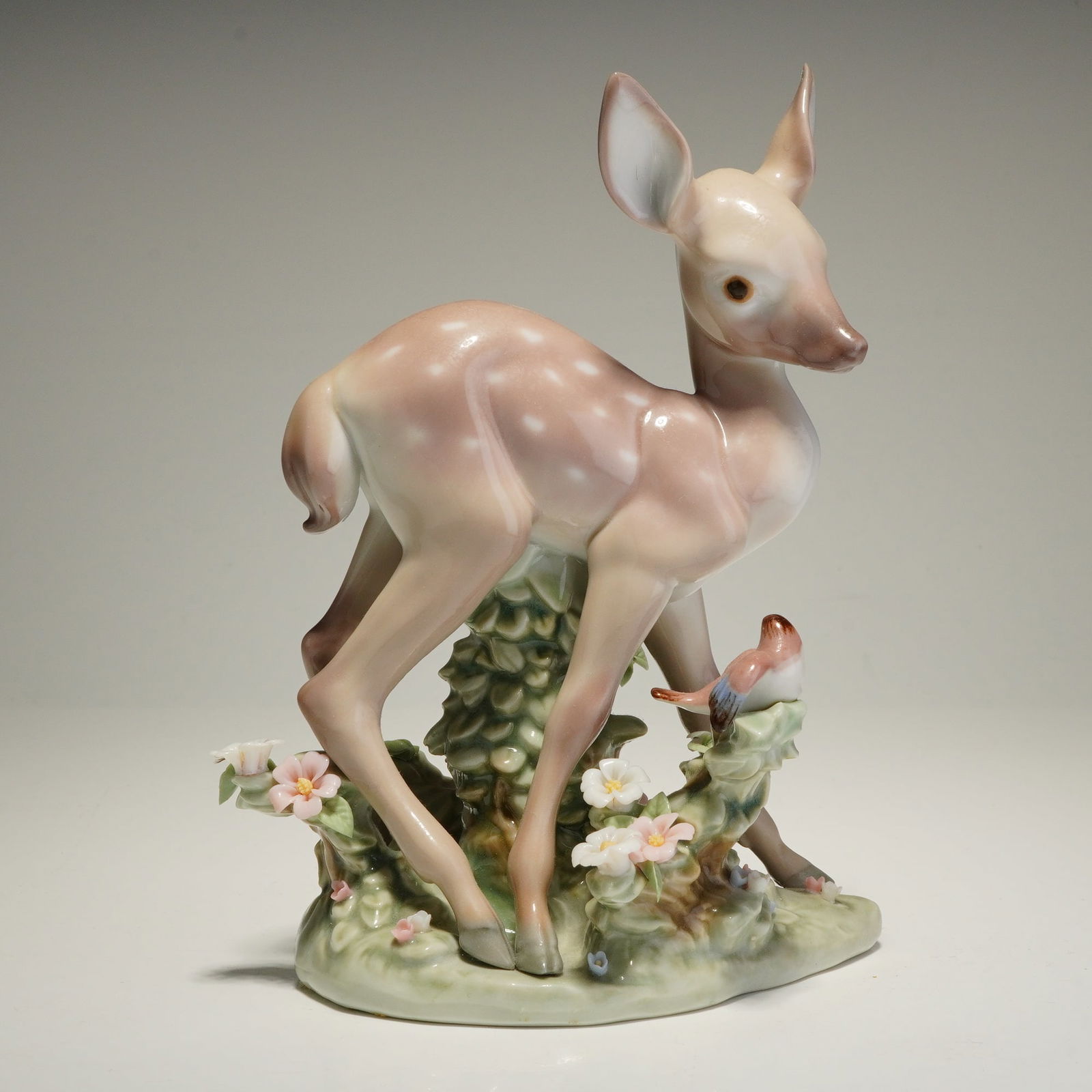 Lladro Porcelain Figurine, A Faun and a Friend 01005674: This glazed porcelain sculpture depicts a spotted fawn standing on a naturalistic base adorned with applied floral elements and a small, perched bird. The piece is hand-finished in a characteristic