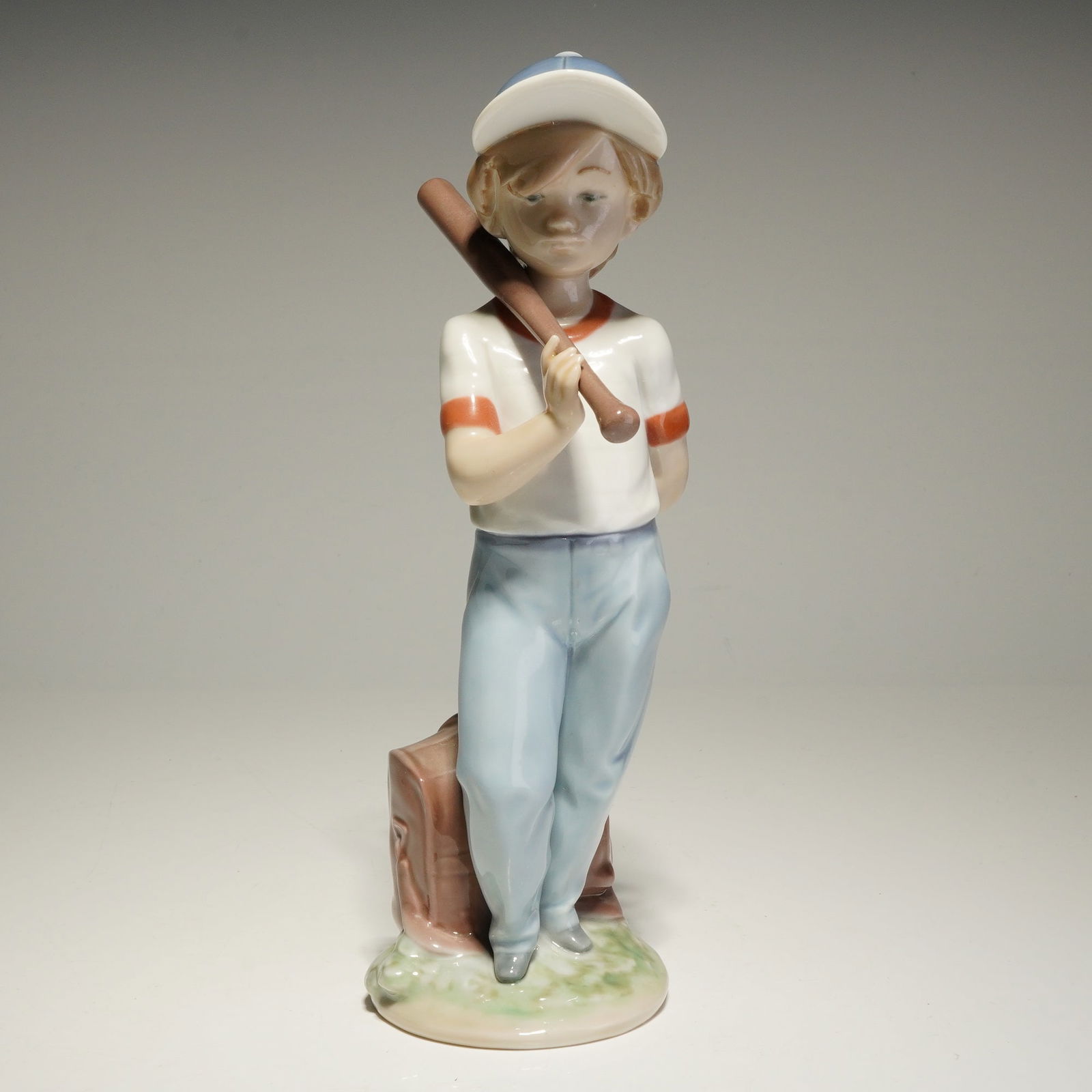 Lladro Porcelain Figurine, Can I Play 01007610: A Lladro porcelain figurine originally produced for the Lladro Collectors Society in 1990. The hand-painted figure depicts a young boy in baseball attire leaning against a brown equipment bag. He hold