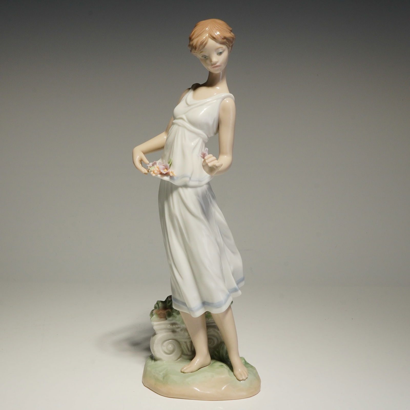 Lladro Porcelain Figurine, Flowers for a Goddess 01007709: A Lladro porcelain figurine from the Privilege collection. The piece depicts a young woman in a sleeveless white dress accented with light blue borders at the waist and hem. She is posed barefoot on a