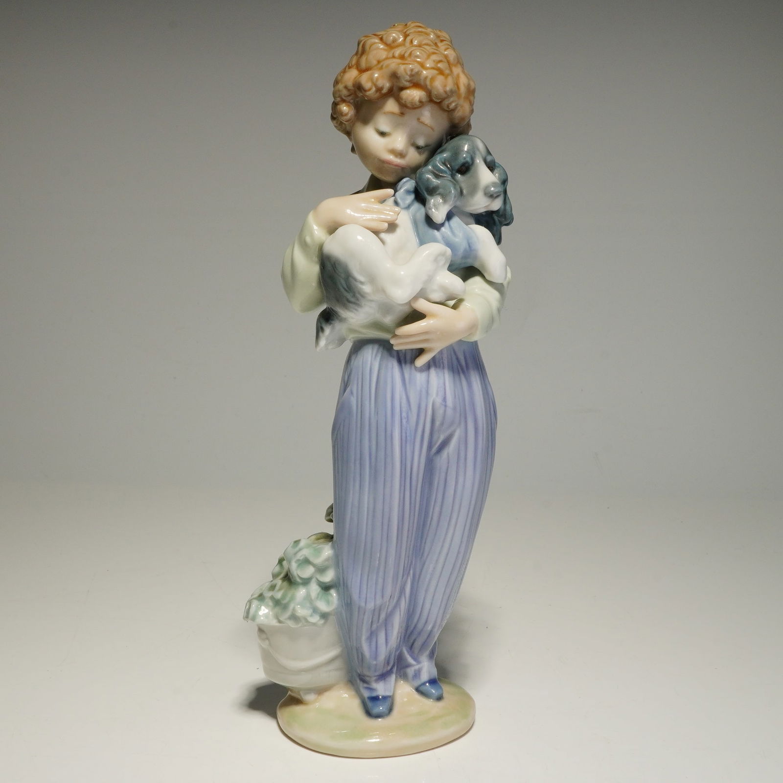 Lladro Porcelain Figurine, My Buddy 01007609: A Lladro glazed porcelain figurine issued for the 1989 Collectors Society. The sculpture depicts a young boy with curly brown hair holding a spaniel puppy. The figure is dressed in lavender-striped tr