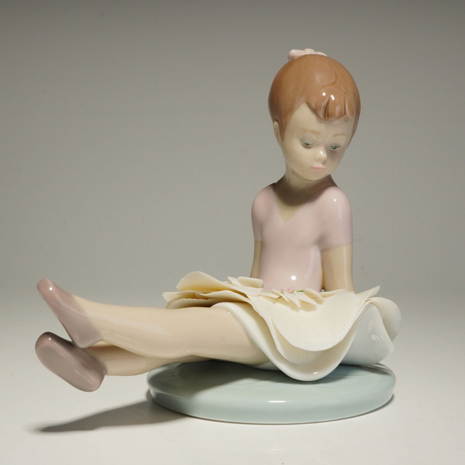 Lladro Porcelain Figurine, Rosy Posey 01006690: A Lladro Rosy Posey glazed porcelain figurine depicting a young girl in ballet attire seated with her legs extended forward. The figure is styled with a pale pink leotard and a voluminous white skirt