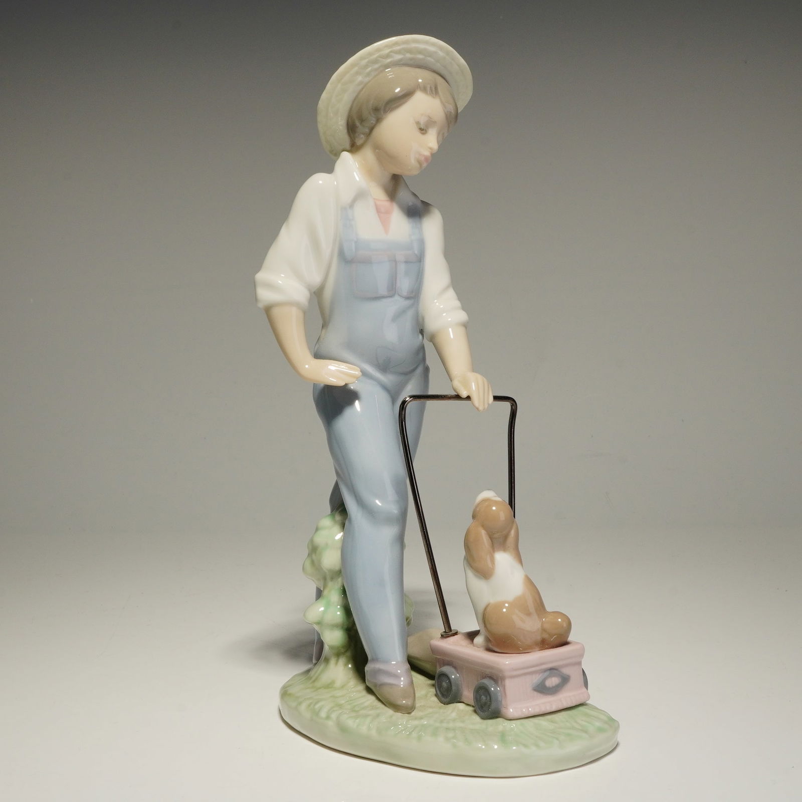 Lladro Porcelain Figurine, Saturday's Child 01006021: This Lladro porcelain figurine is a glazed figure depicting a young boy in light blue overalls and a straw hat. The boy is shown pushing a small pink wagon that contains a seated brown and white dog.